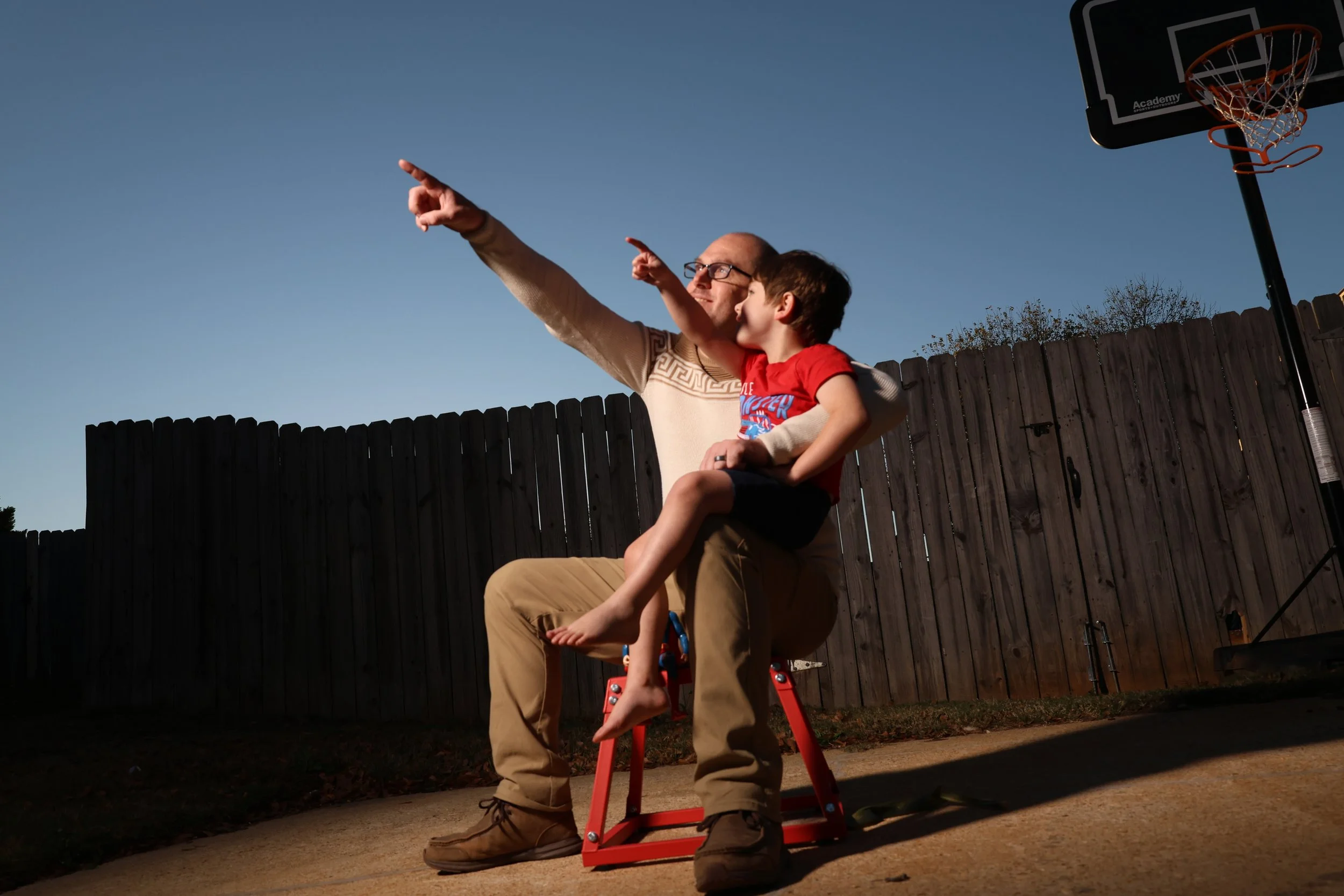 An adult and a child sitting together on a red chair outdoors, pointing at something in the sky during sunset.