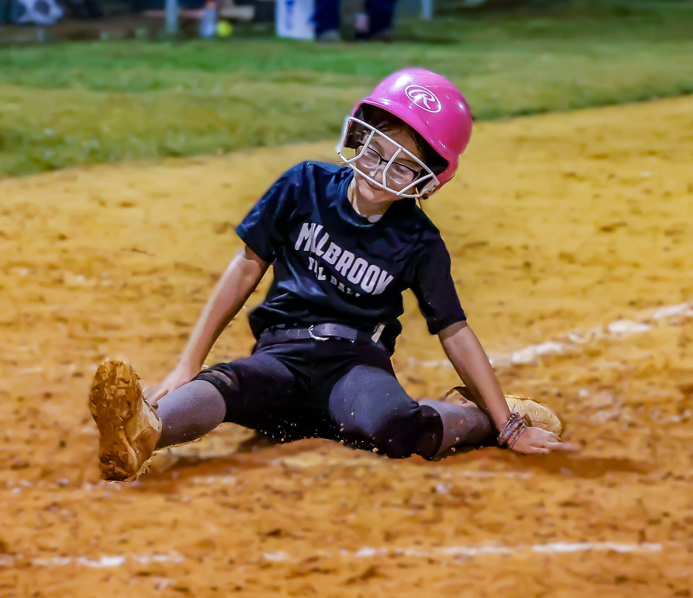 A young softball player in a pink helmet and dark jersey sliding on the dirt in a softball field.