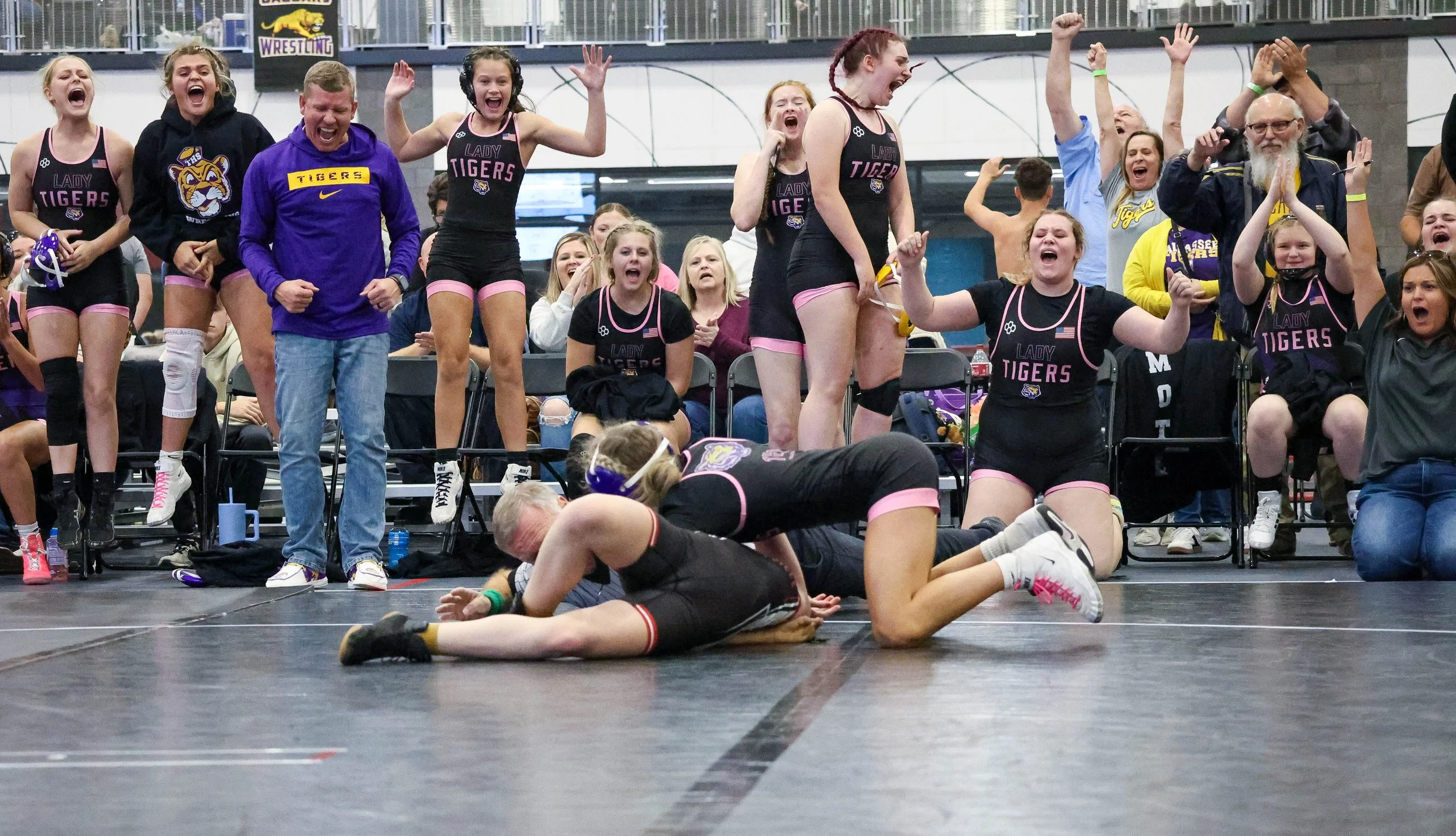 Wrestlers in black and pink uniforms celebrate a wrestling match after winning a match. Fans and teammates cheer and applaud in the background.