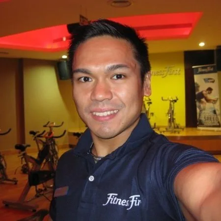 A man taking a selfie in a gym, smiling, wearing a navy blue polo shirt with 'Fitness First' logo, with fitness equipment and a yellow wall in the background.