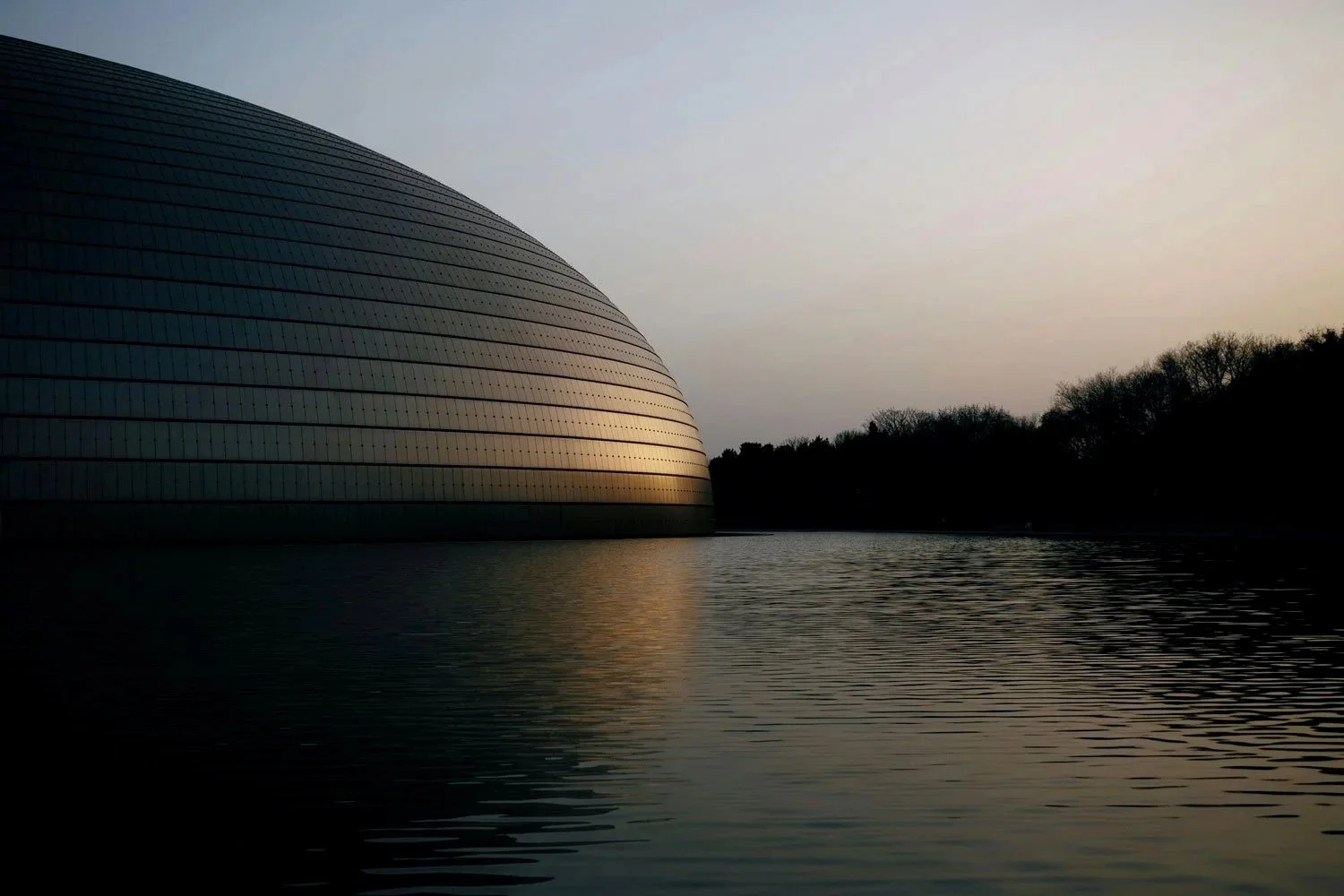 A large, modern building with a rounded, shell-like shape at the water's edge during sunset, with trees in the background and calm water reflecting the building.