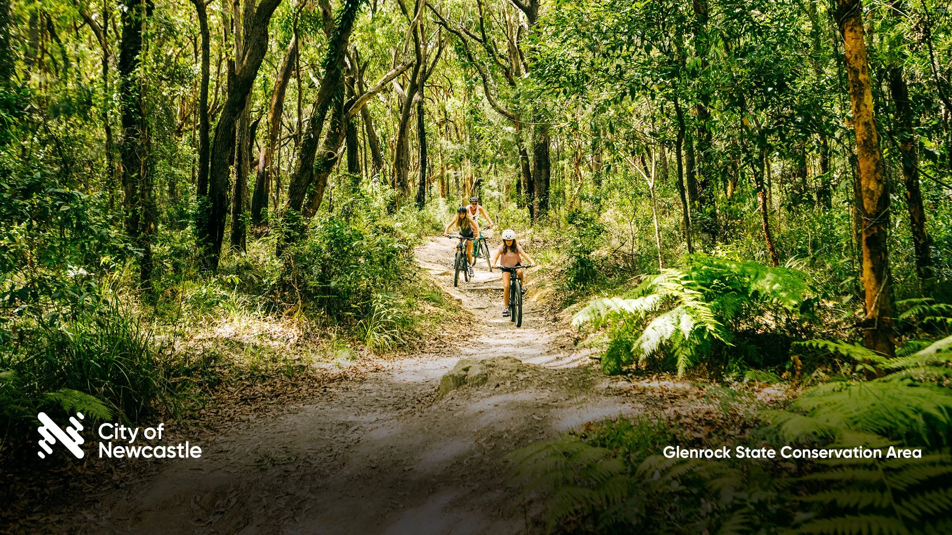 Four people riding bikes on a dirt trail through a lush, green forest in Glenrock State Conservation Area near Newcastle.