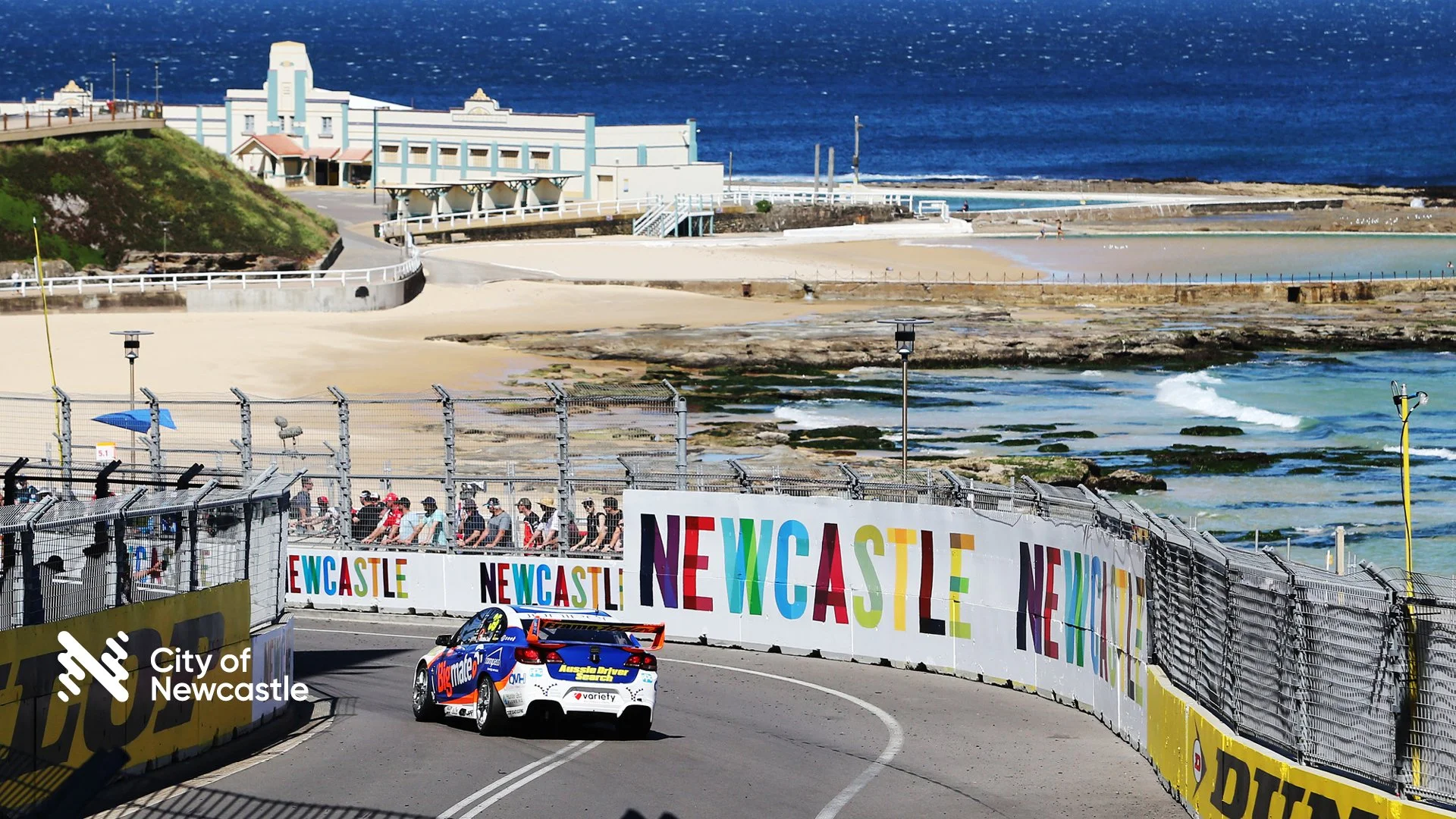 A race car on a track near the beach, with spectators behind fencing, and colorful NSWCASTE banners. The ocean and sandy beaches are visible in the background.