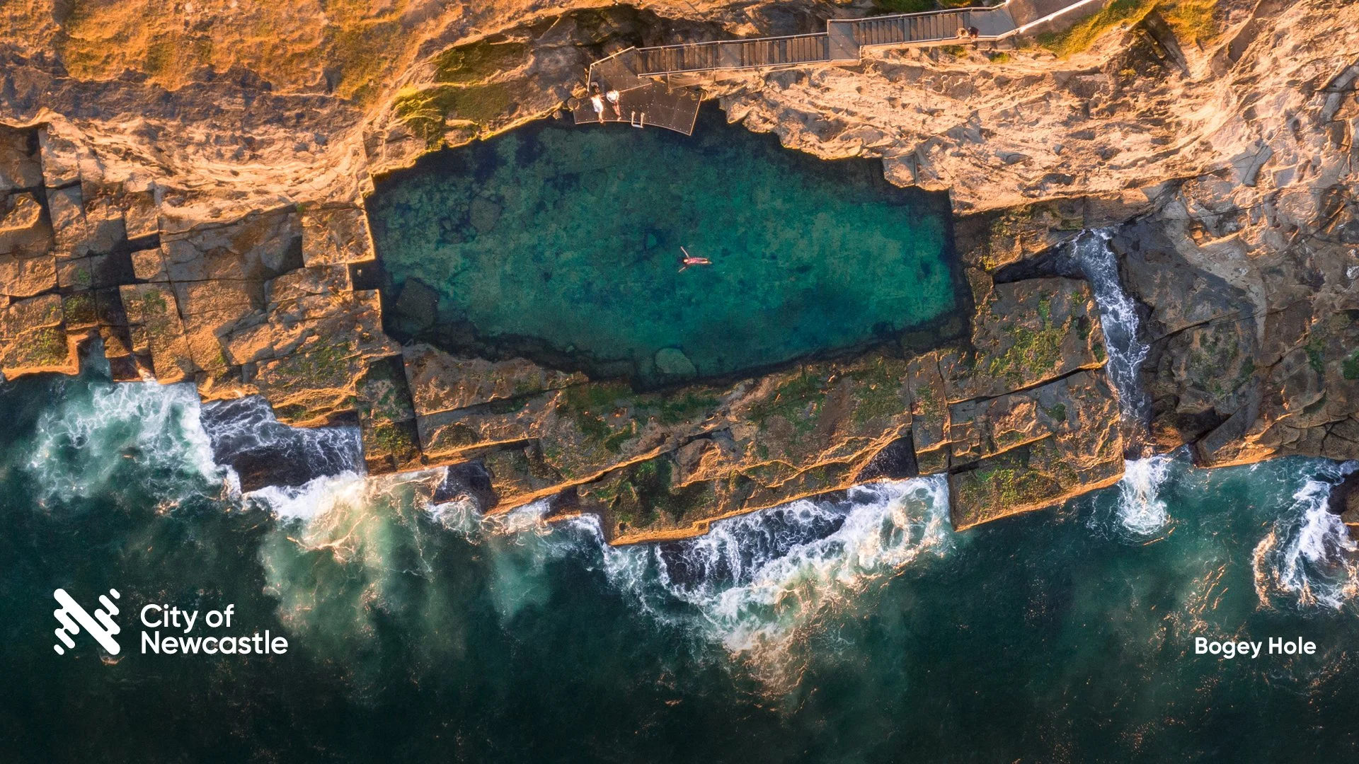 An aerial view of Bogey Hole, a rock pool along the coast in Newcastle, Australia, with waves crashing against the rocks and a person paddleboarding in the water.