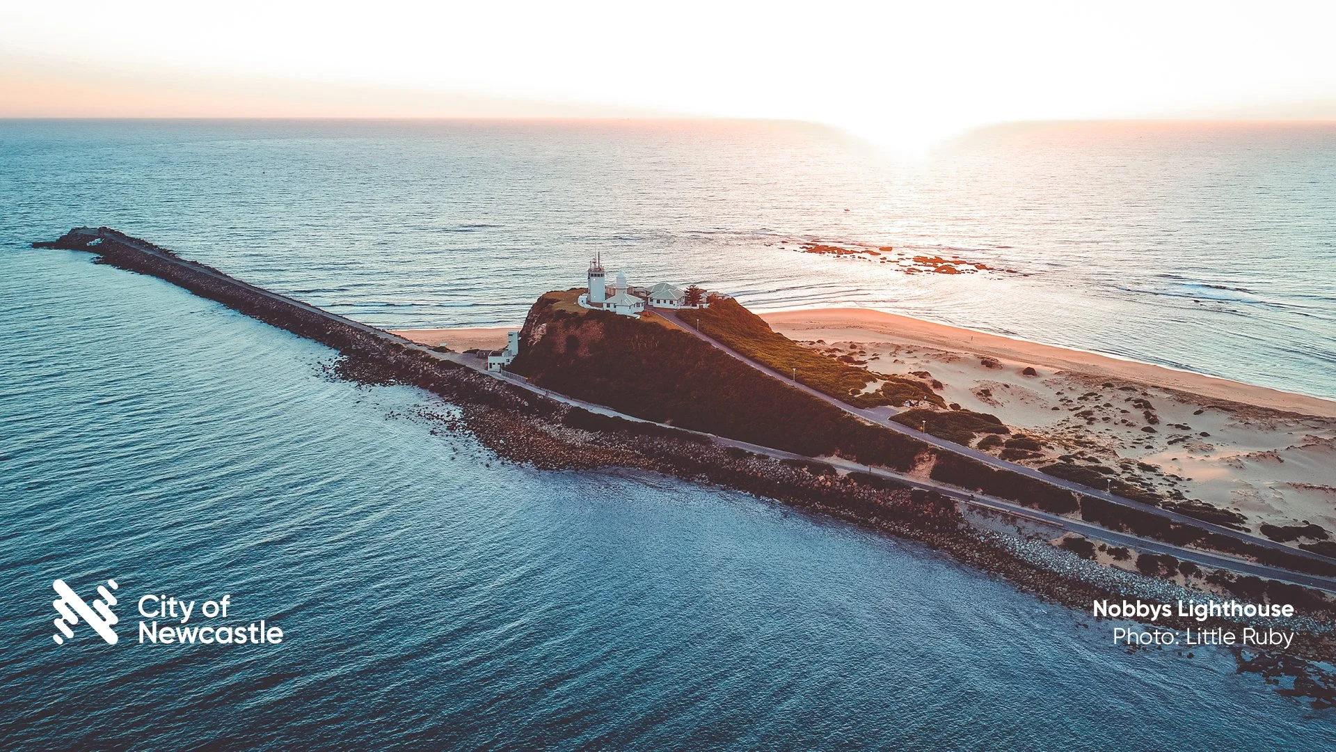 An aerial view of Nobbys Lighthouse on a small headland with sandy beaches and rocky areas, extending into the ocean at sunset, with a clear sky and gentle waves.
