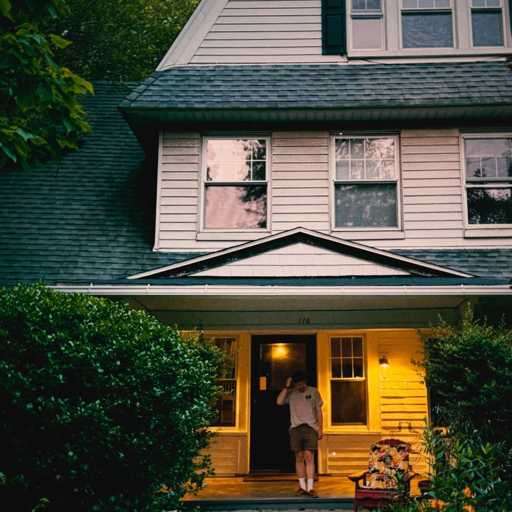 Front porch of a house at twilight with two people standing near the door. One person is standing outside on the porch, and another is inside the house near the door. The house has a light yellow exterior with a dark front door and surrounding windows. There are lush green bushes on both sides of the porch.