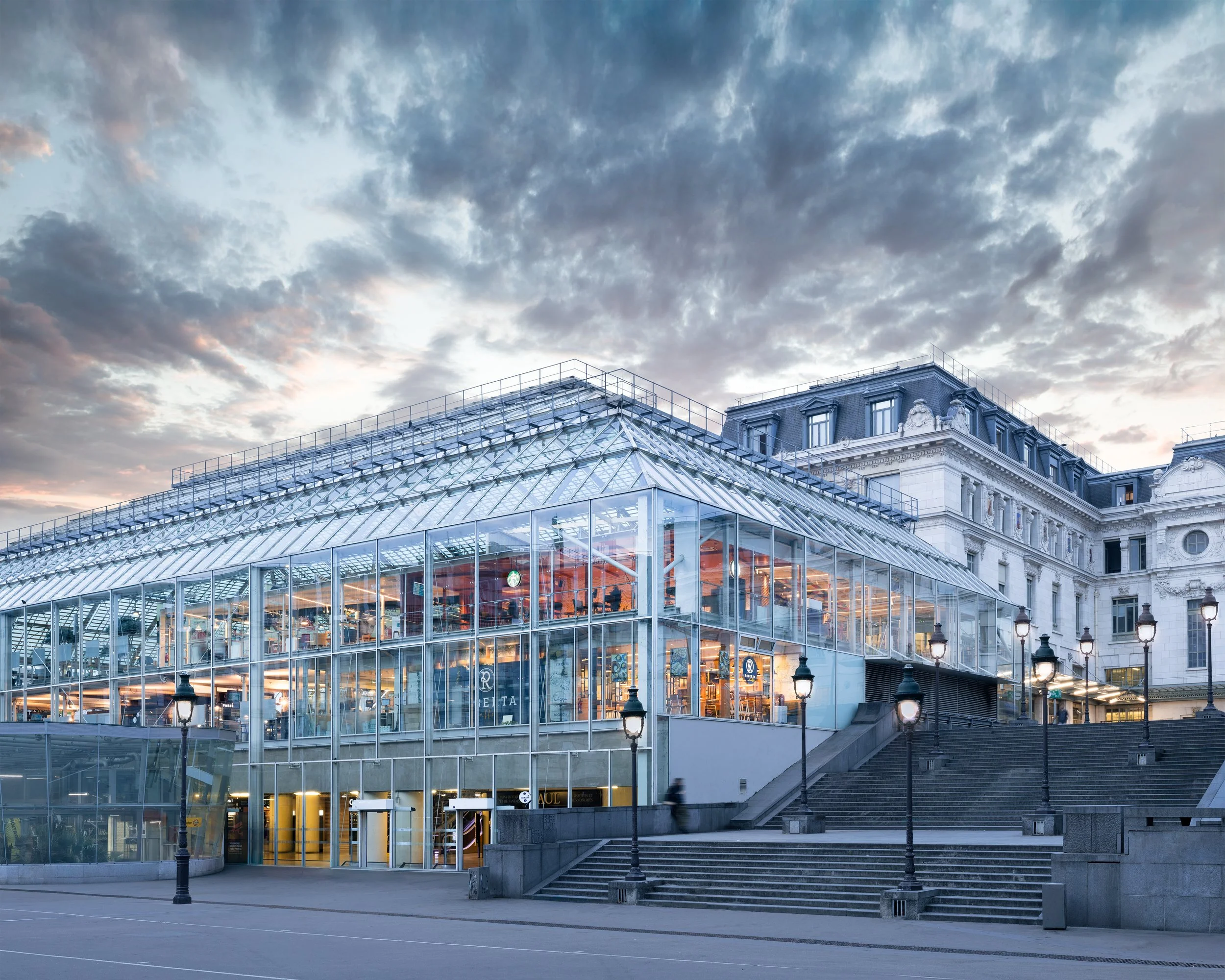 Modern multi-story glass building with stairs and lampposts outside, classic architecture in background, cloudy sky at dusk.
