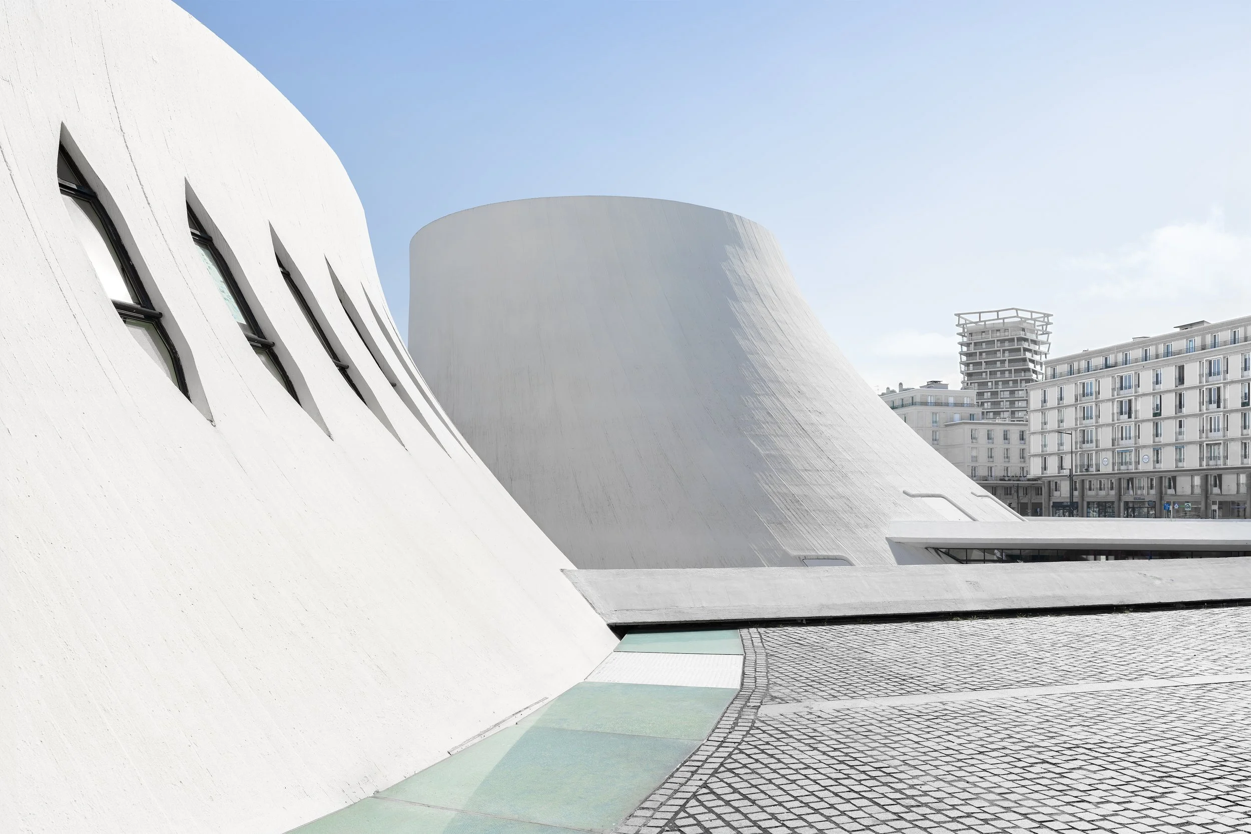 Modern white architectural building with curved walls, small elongated windows, and a cityscape background.