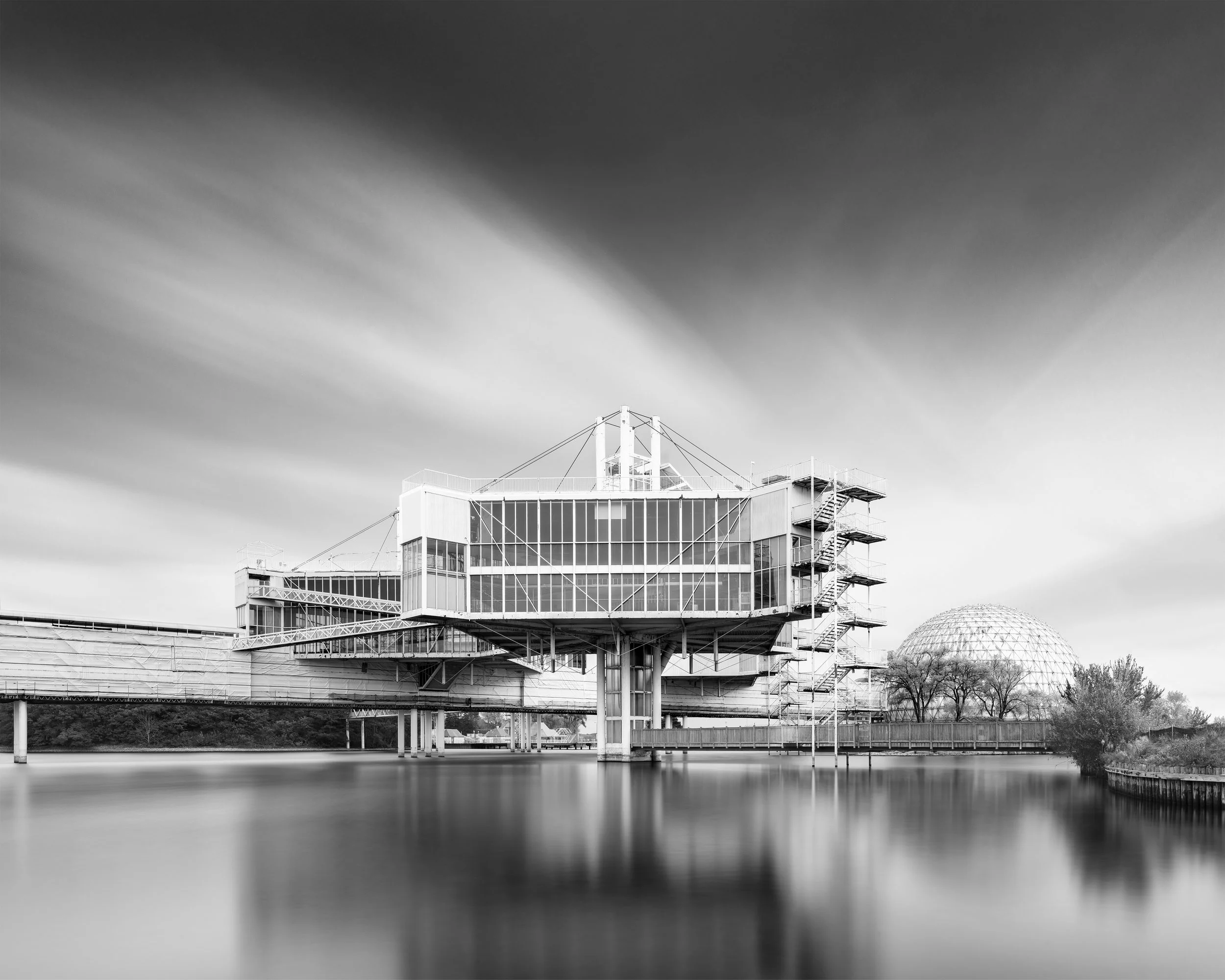 A black and white photo of a modern architectural building with large glass windows, situated over water, and a dome structure in the background.