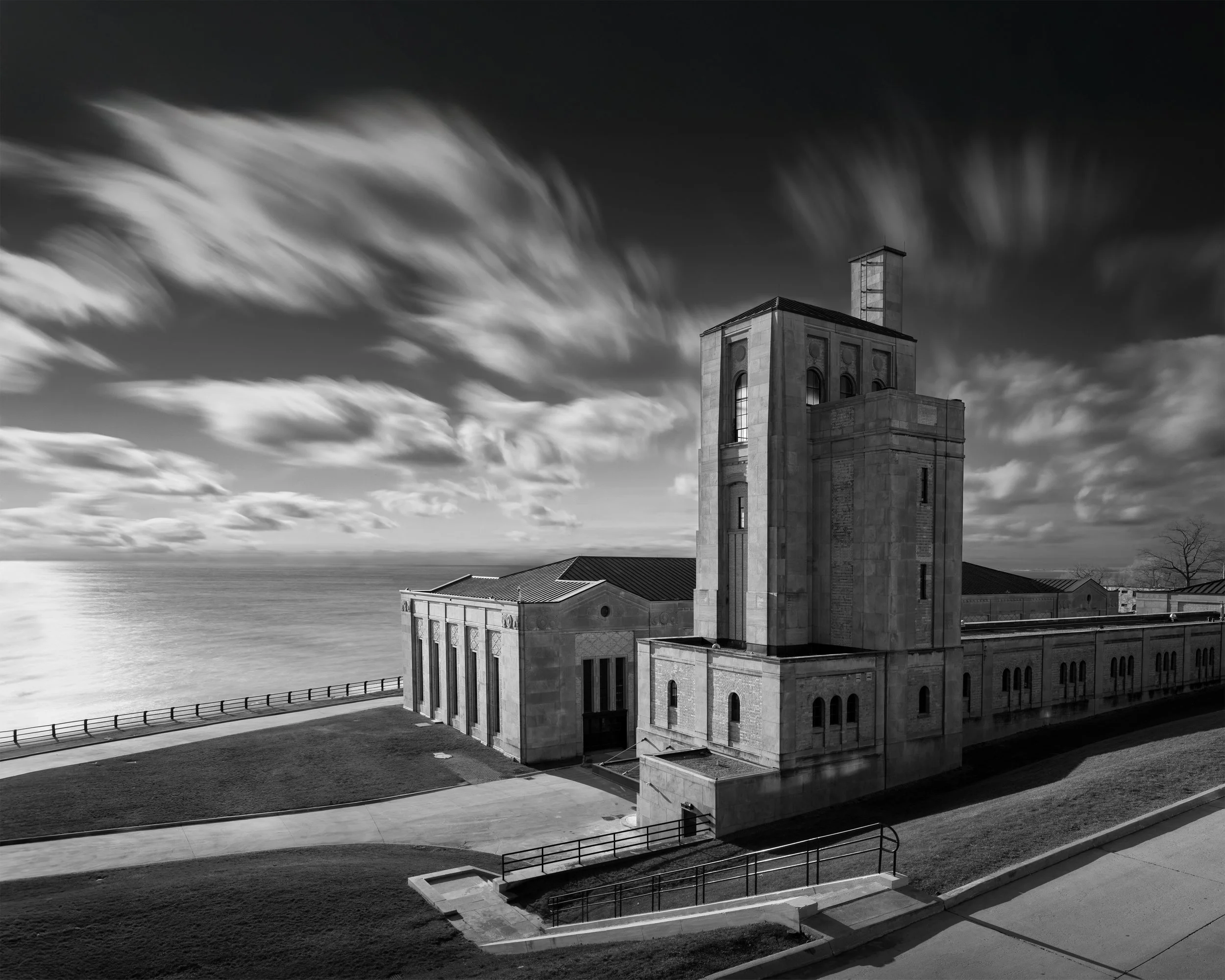 Black and white photo of a large, historic building with a tall clock tower by the water with cloudy skies.