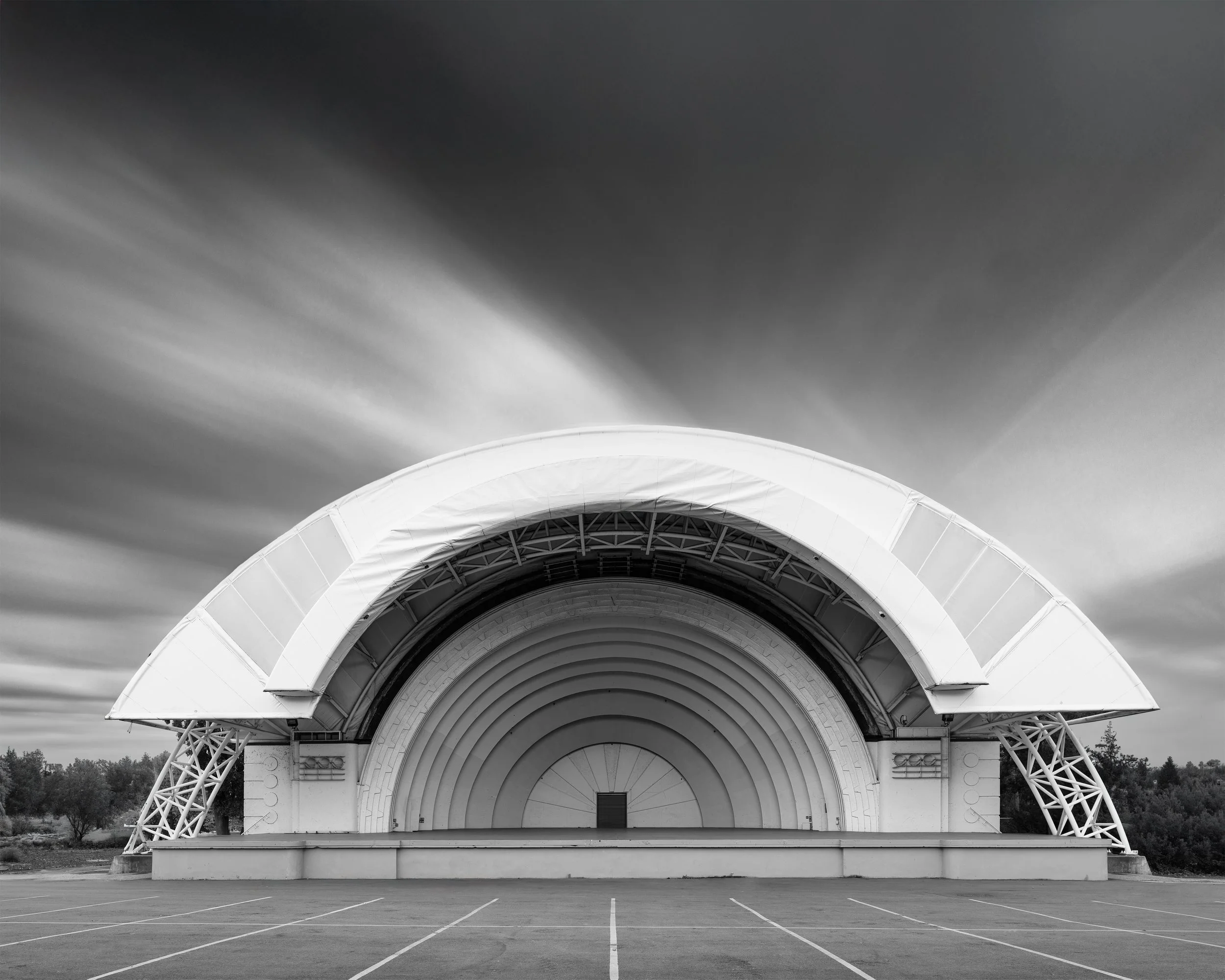 Black-and-white photo of a large outdoor band shell or amphitheater with a curved roof and open stage, empty parking lot in the foreground, trees in the background, and clouds in the sky.