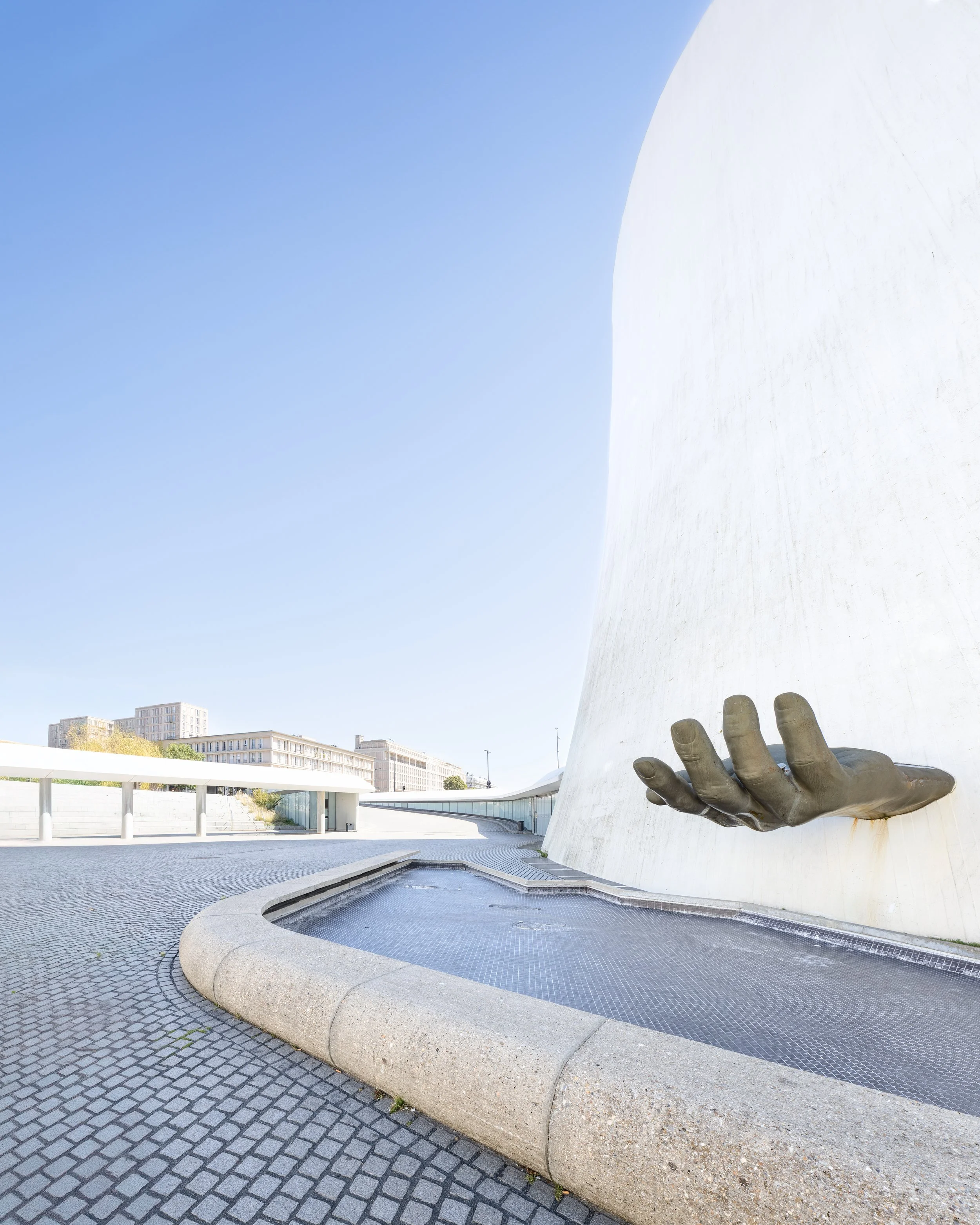 A large white modern building with a prominent bronze sculpture of a hand emerging from the wall, situated near a fountain, on a sunny day with clear blue sky, in an urban area.