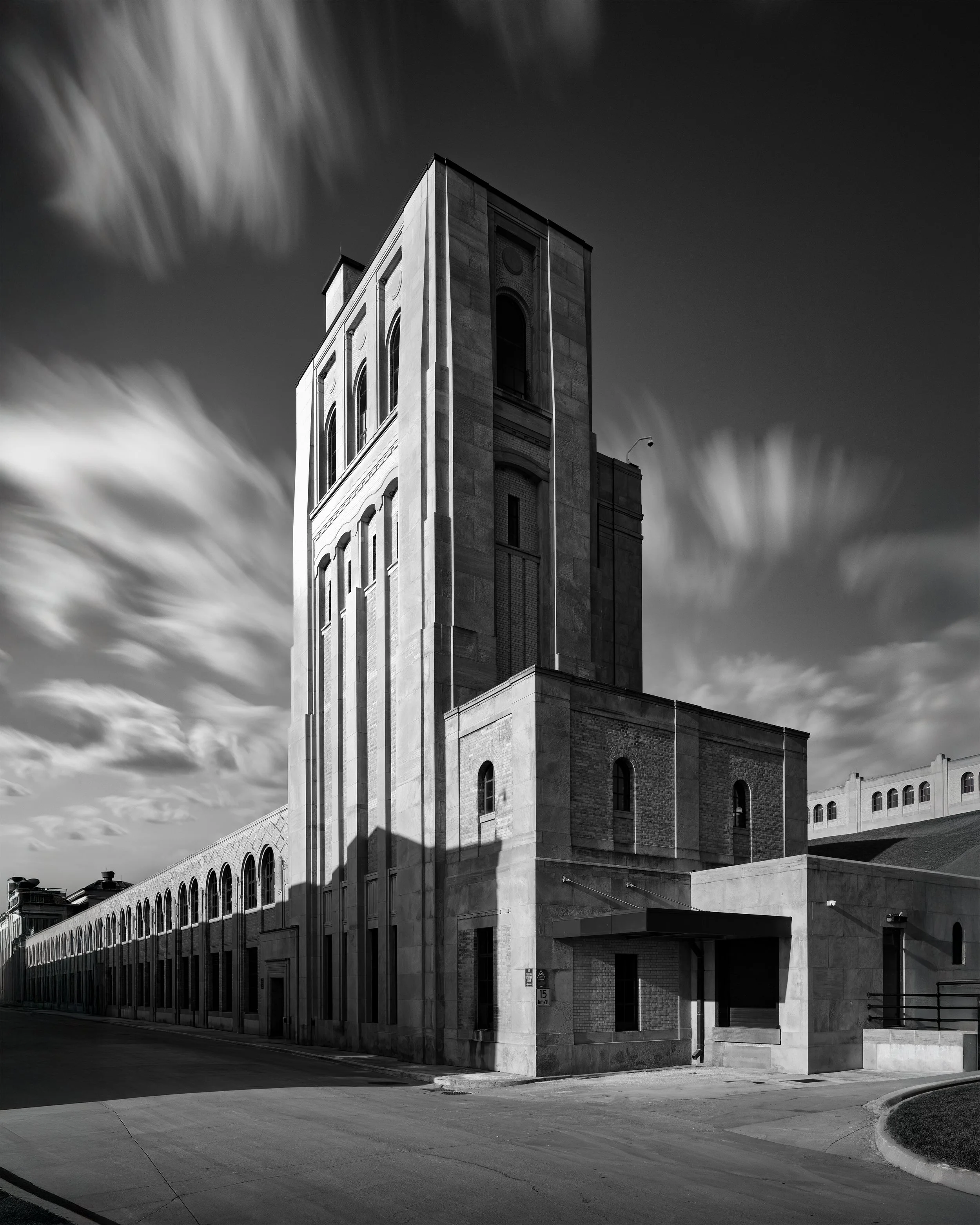 Black and white photo of a tall, modern building with arched windows and a long row of arches at the base, under a sky with fast-moving clouds.
