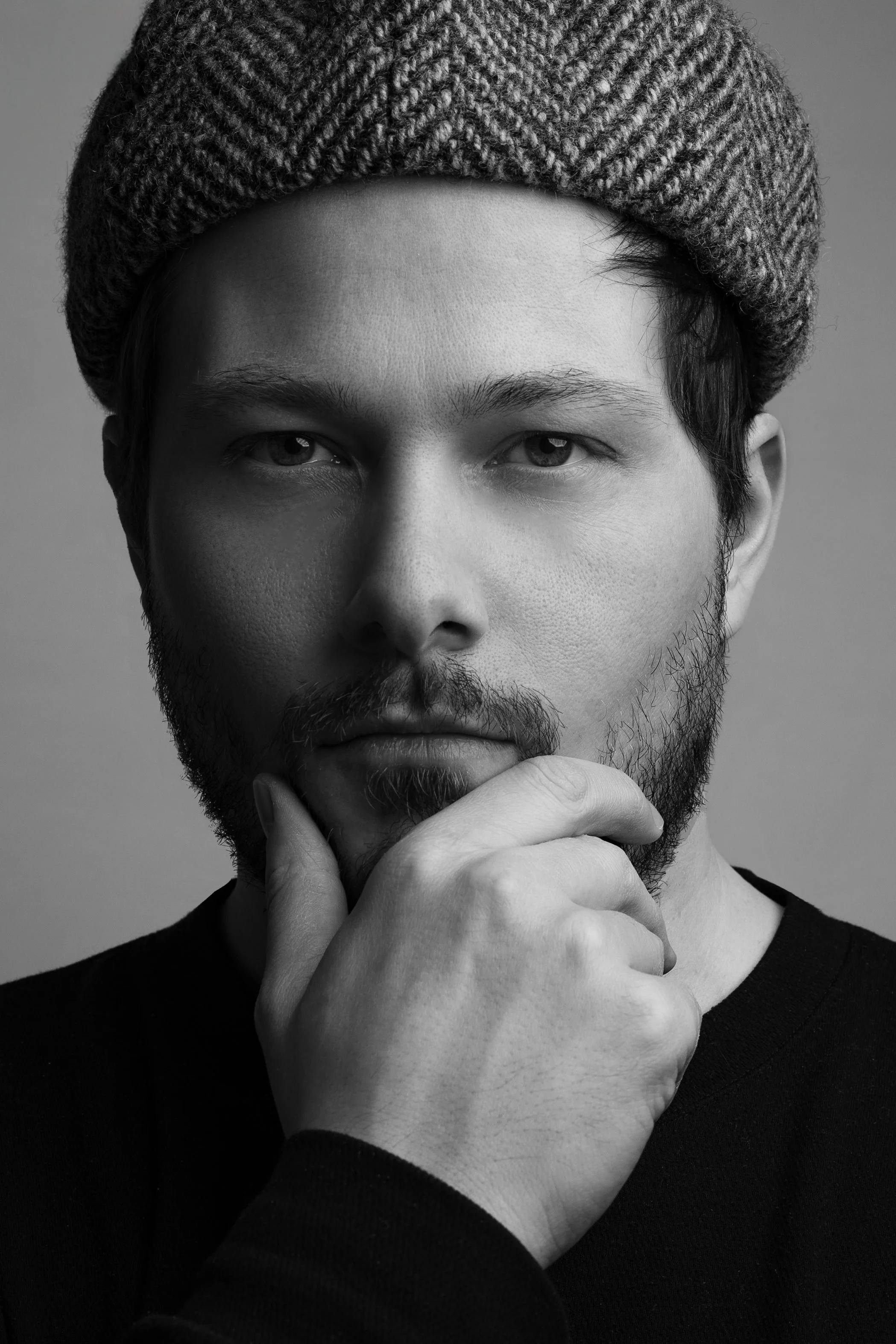 Black and white portrait of a man wearing a herringbone patterned hat, with his hand on his chin, looking thoughtfully at the camera.