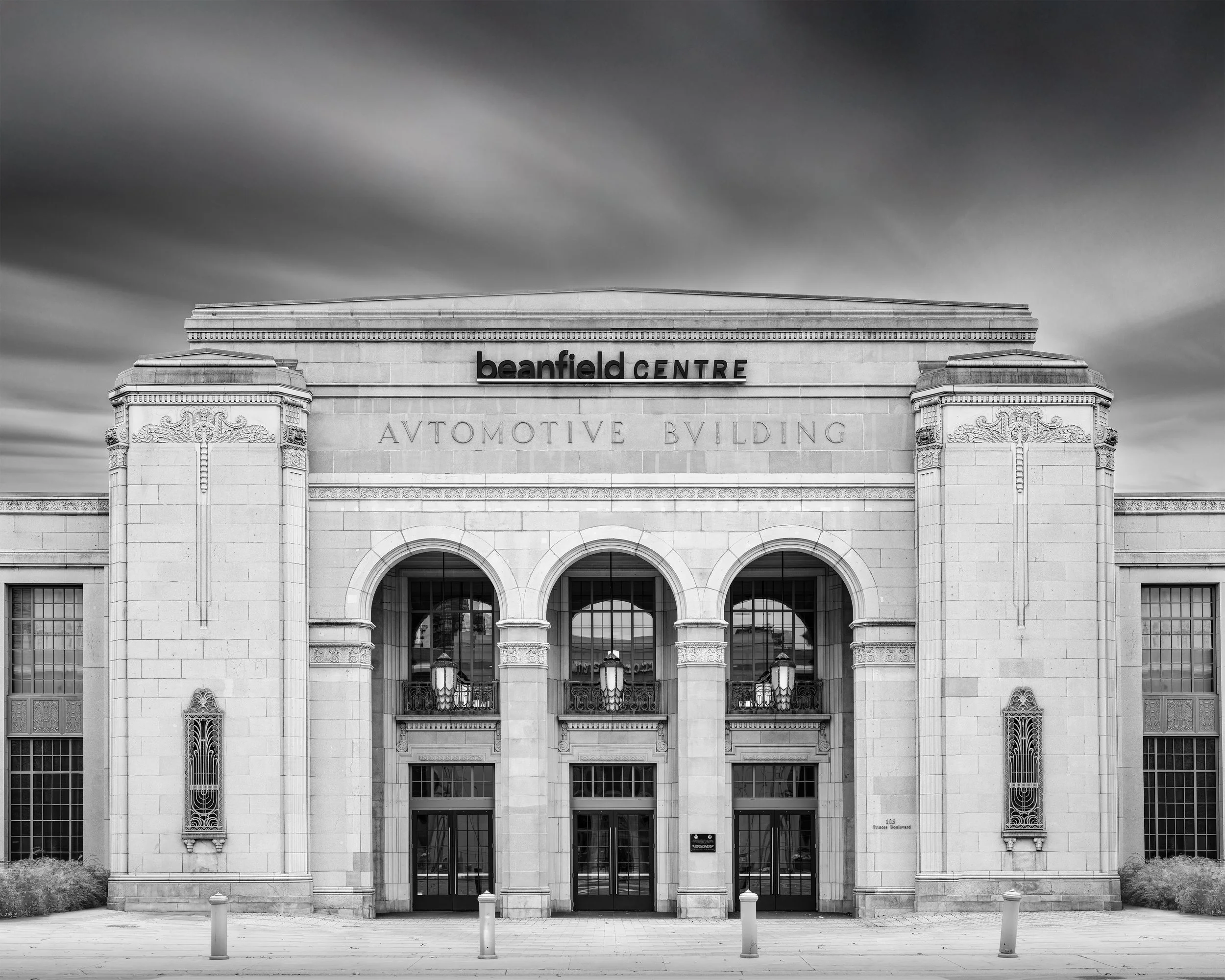 Black and white photo of the Beanfied Centre's front facade, featuring three large arched windows and intricate architectural details, with a cloudy sky overhead.