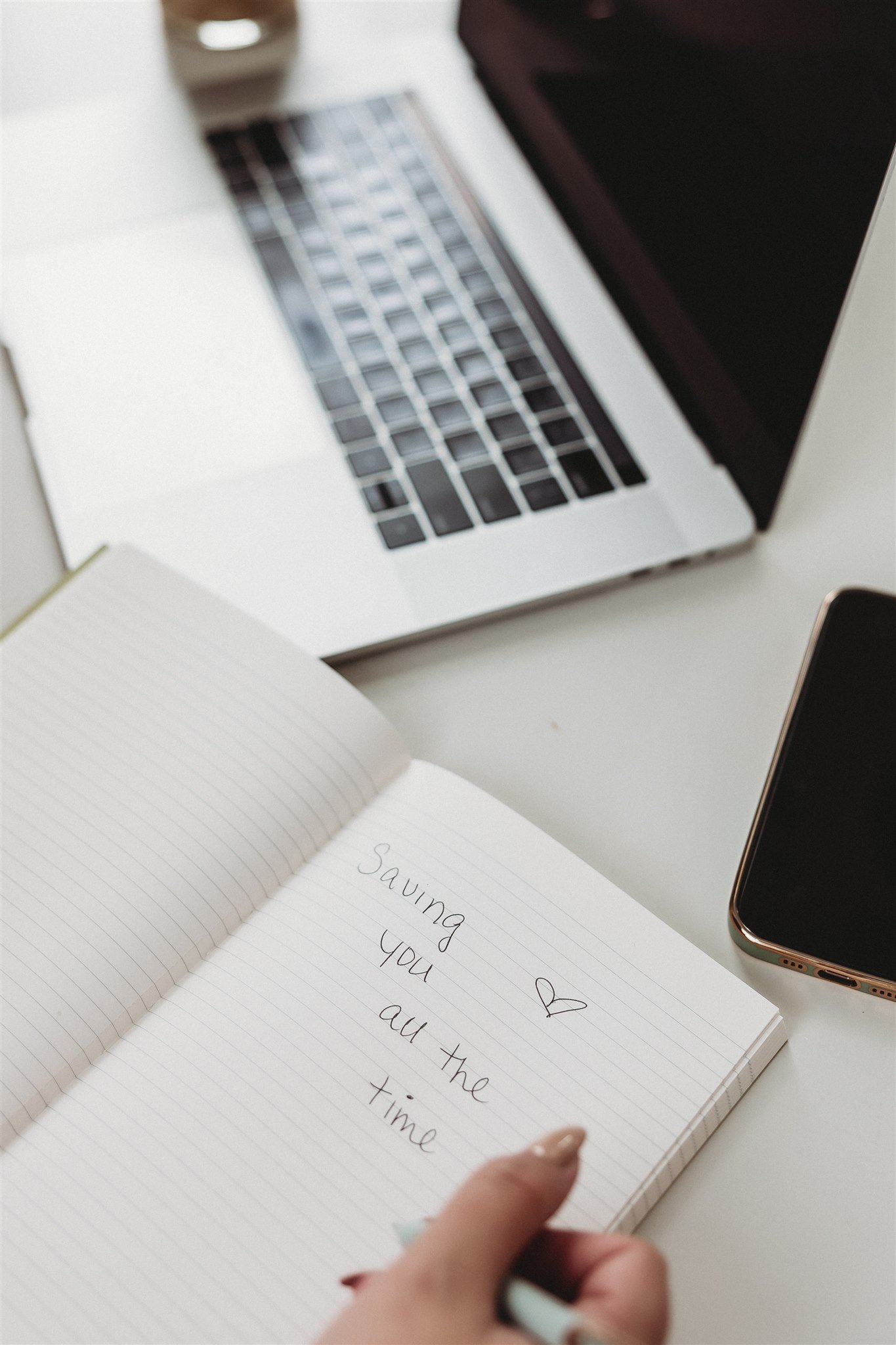 Open notebook with handwritten text that reads 'Saving you ❤️ all the time', on a white desk with a laptop and a smartphone.
