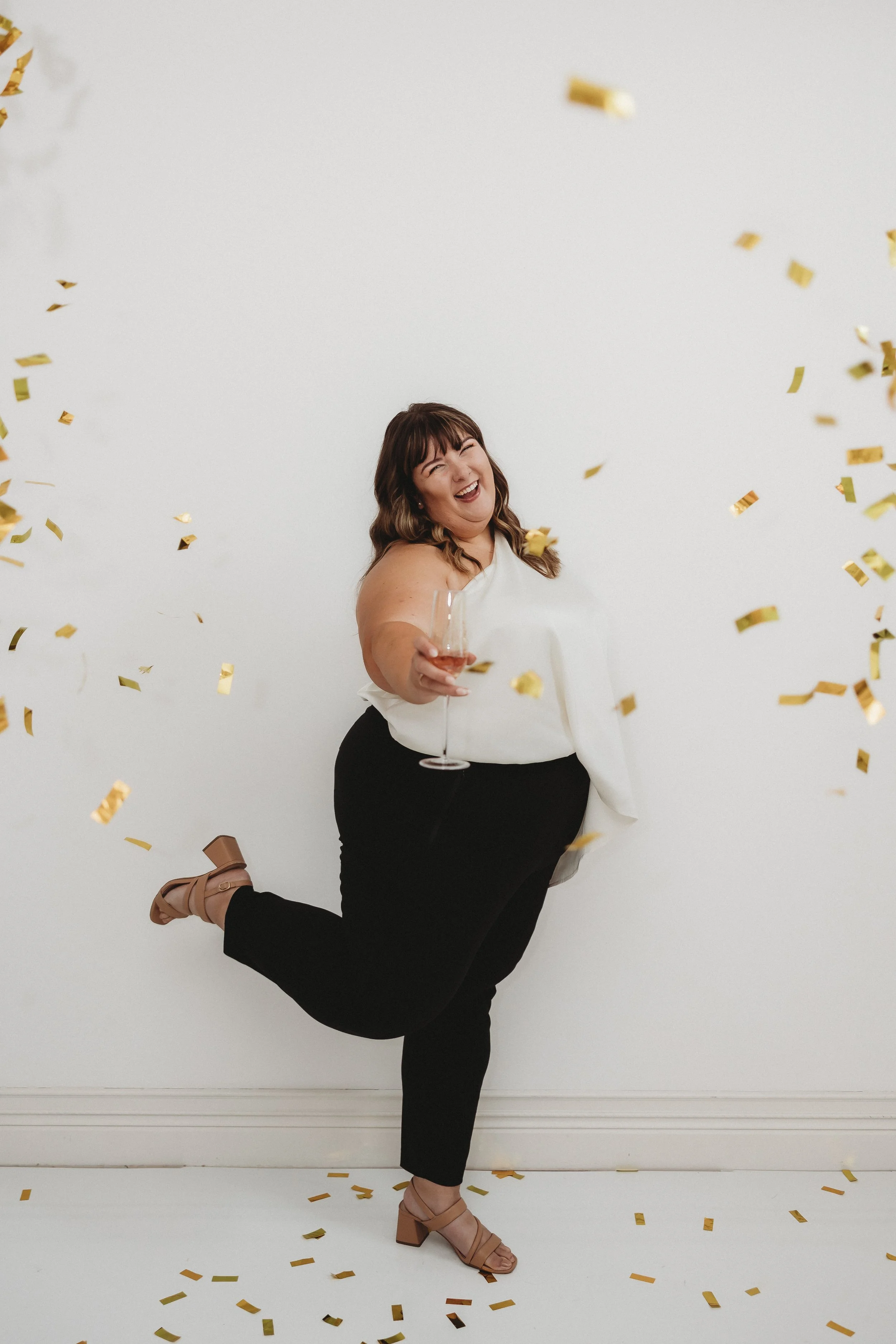 Bre Henderson, owner of Tended Sprout, smiling and celebrating with confetti, holding a glass of champagne, standing against a white background.
