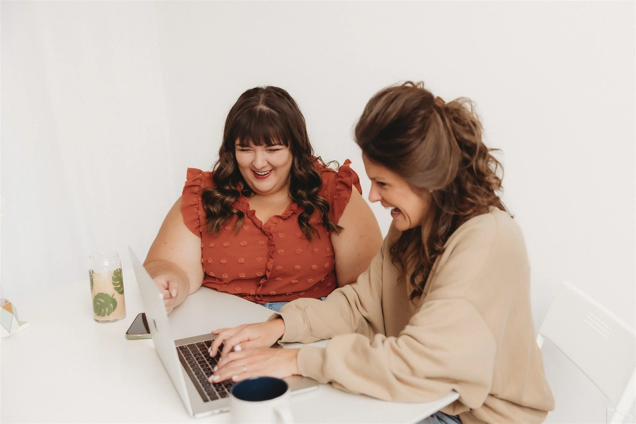 Two women sitting at a white table, smiling and looking at a laptop together, engaged in a cheerful conversation.