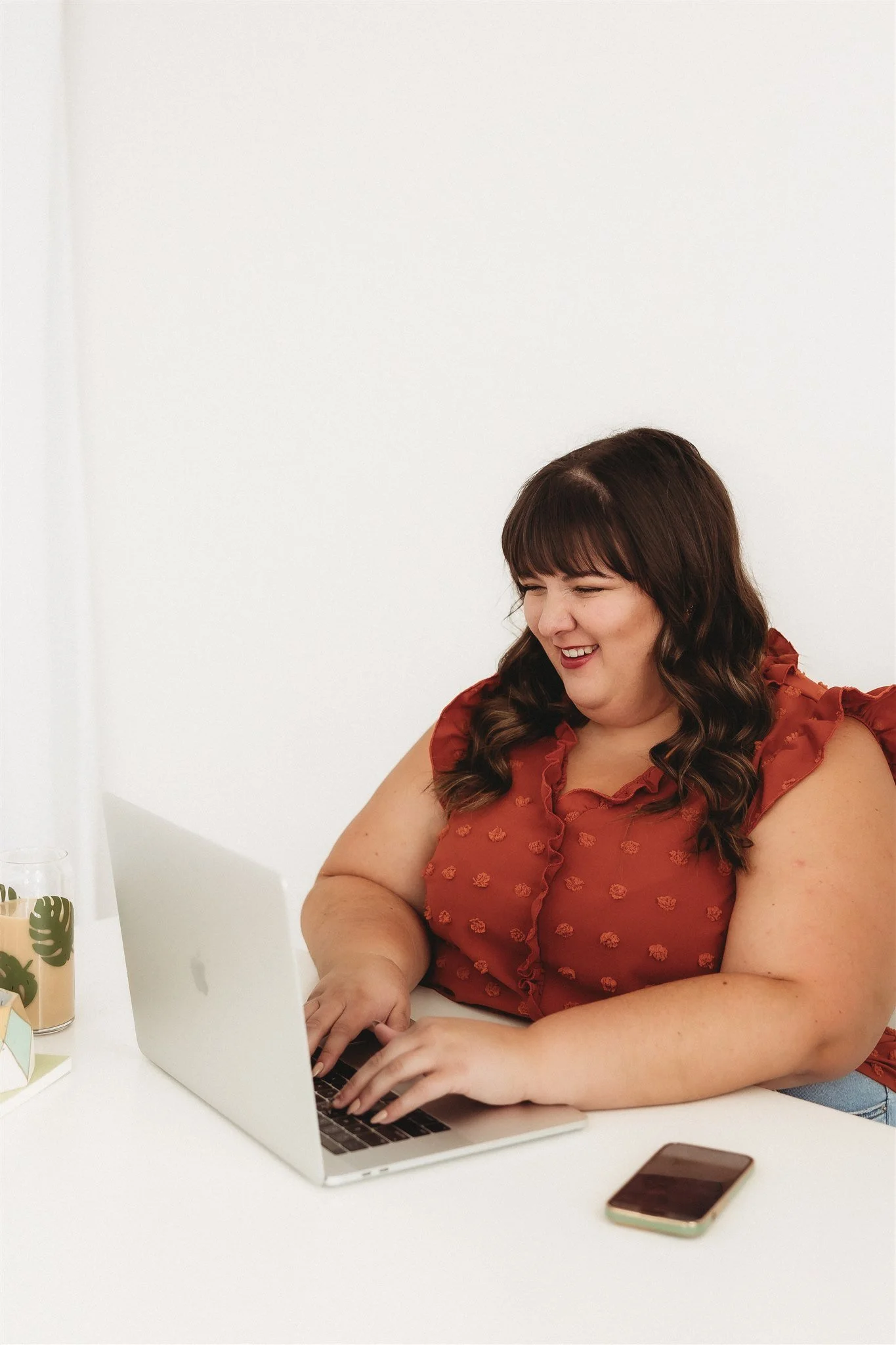 A woman with dark brown hair, wearing a rust-colored blouse, sitting at a white desk, smiling while using a silver laptop. A smartphone is placed on the desk beside her, and a decorative plant with large green leaves is also on the desk.