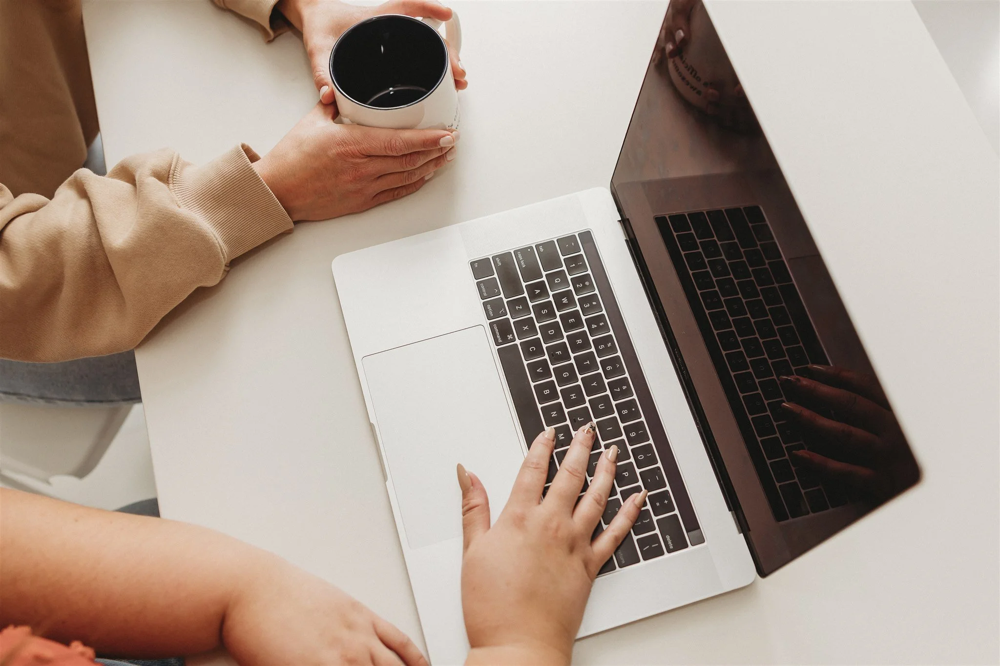 Person working on a laptop at a white table, holding a cup of coffee in one hand and typing with the other, with a child's arm resting on the table.