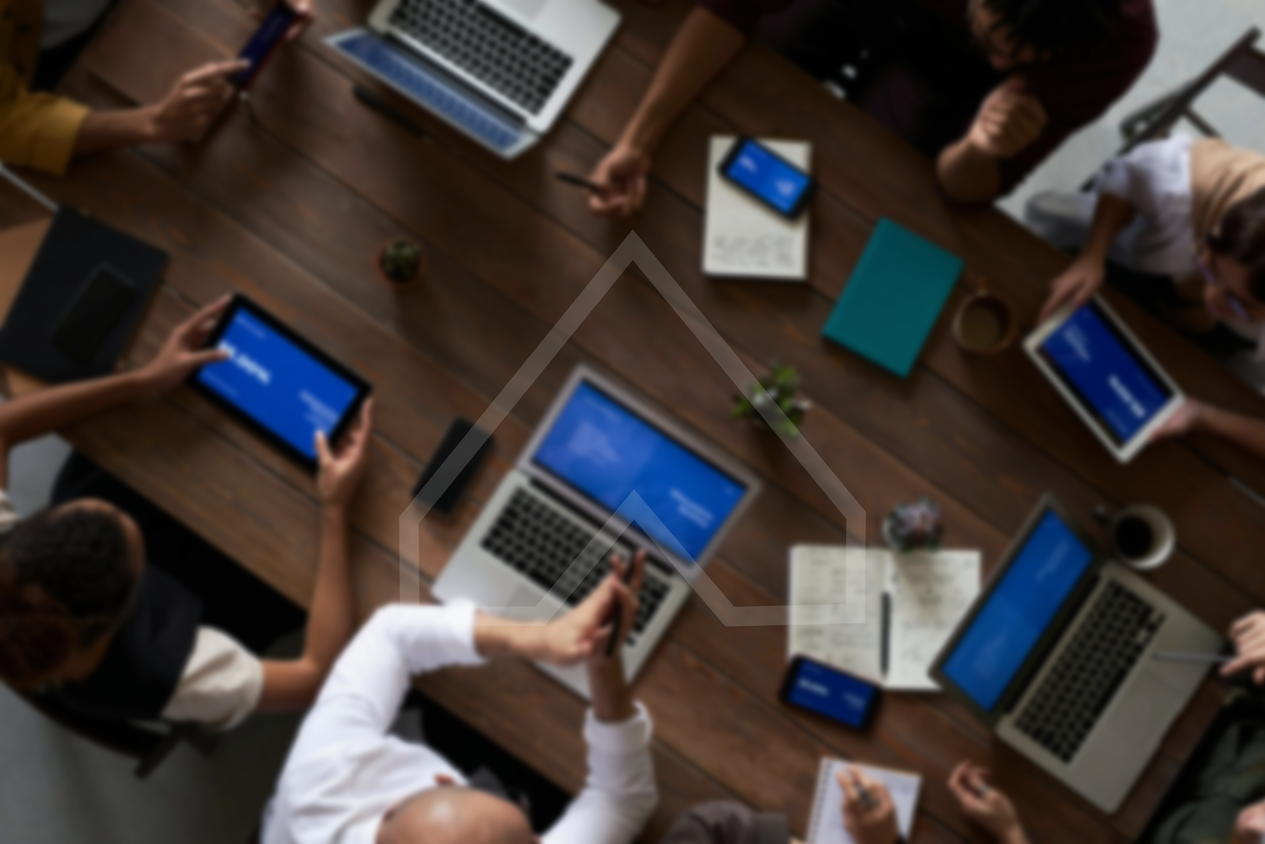 A top-down view of a meeting room with multiple people working on laptops and tablets, some taking notes and others using smartphones, with a wooden table, notebooks, potted plants, and coffee cups.