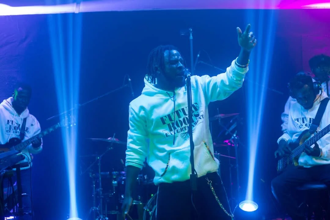 A male singer with dreadlocks wearing a white hoodie with black text, performing on stage with a band under colorful stage lights.