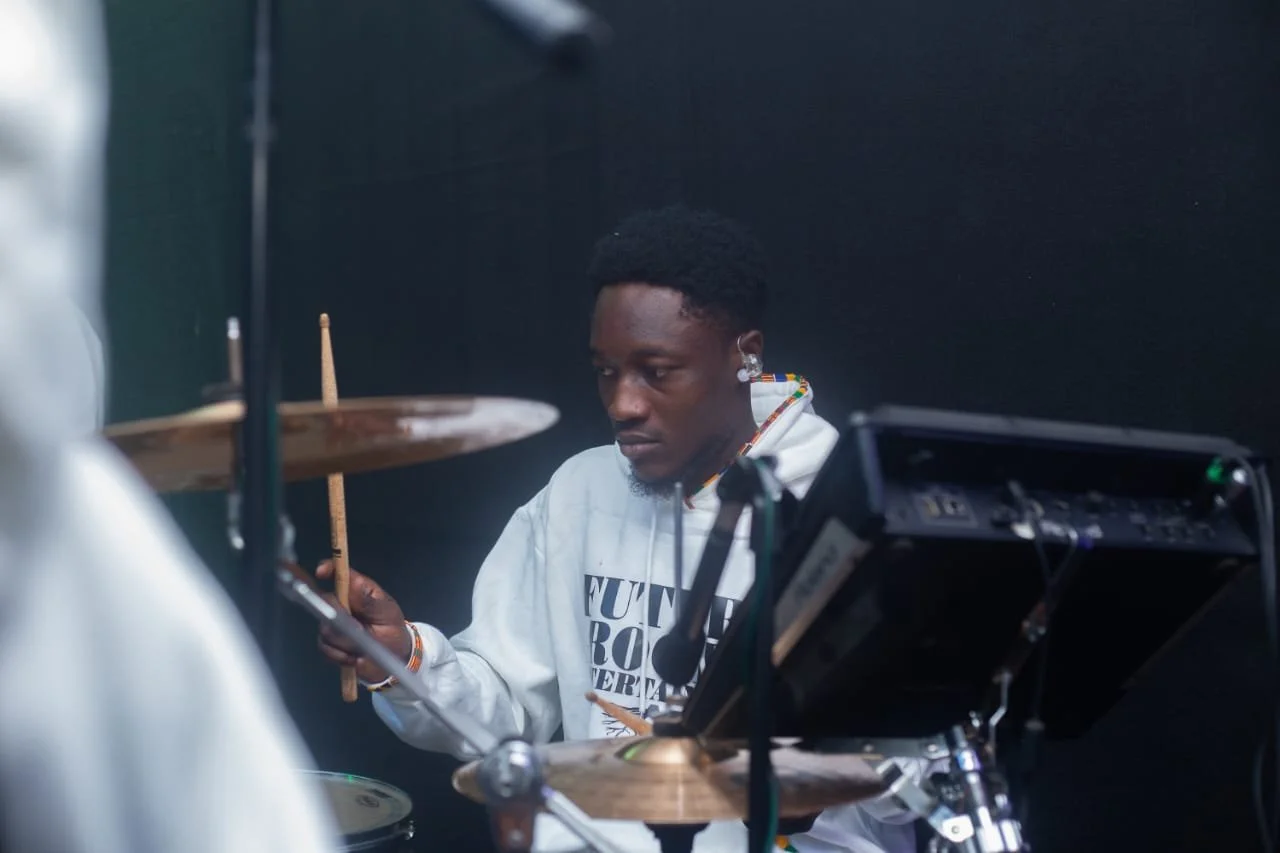 Young man playing drums in a music studio, wearing a white hoodie with text and beaded jewelry, focused on his drumming.