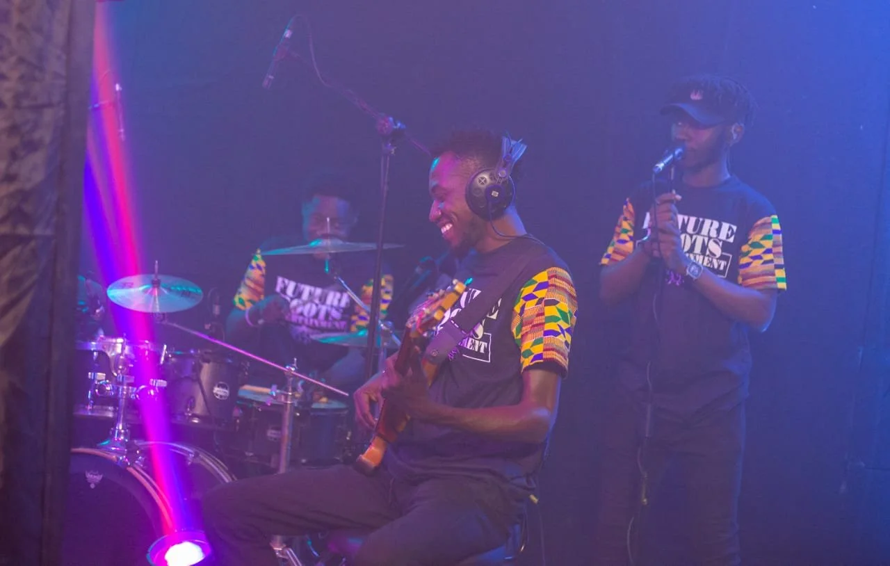 Three musicians performing on stage with colorful lighting. One playing the drums, another playing the guitar, and the third singing into a microphone. All wearing matching shirts with patterned sleeves.