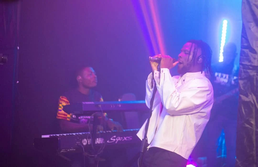 A singer with braided hair wearing a white shirt performs with a microphone in a dimly lit, colorful nightclub. A keyboardist is playing in the background.