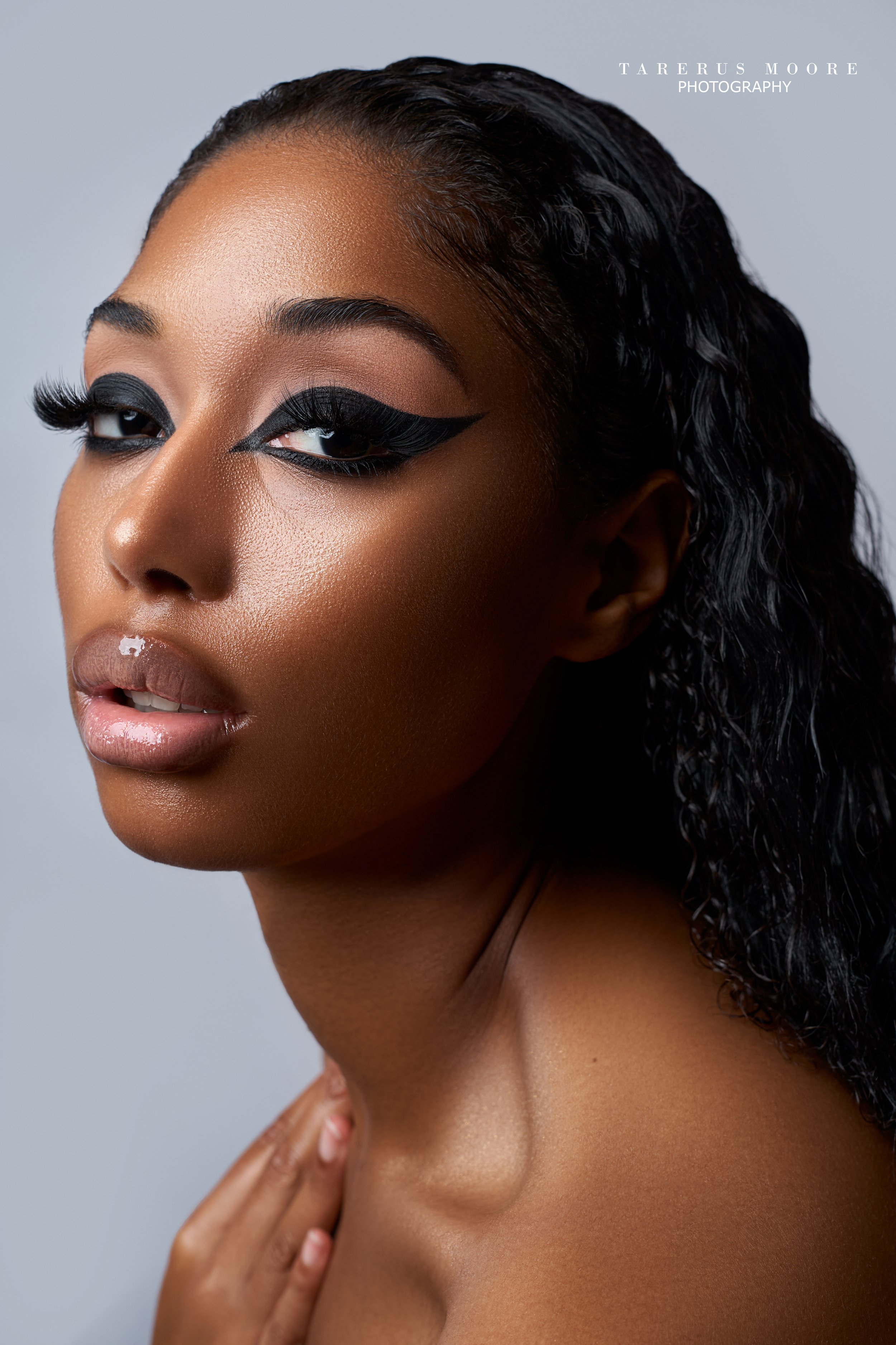 Close-up portrait of a woman with glamorous makeup, including bold black eyeliner, long lashes, and glossy lips, with textured skin and curly black hair, against a plain light gray background.