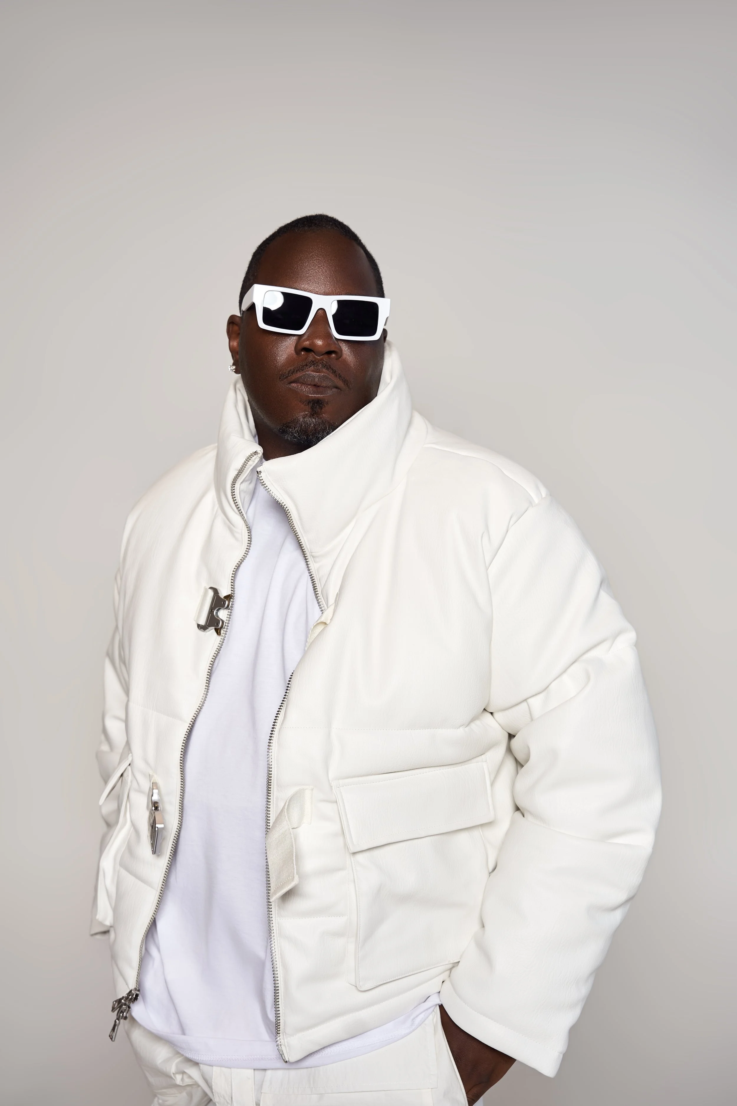 A man wearing white sunglasses, a white high-collared jacket, and a white shirt, posing against a plain light background.
