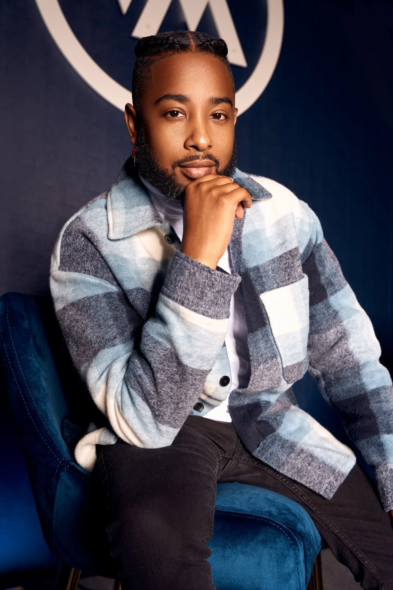 A man with a beard and styled hair, wearing a checkered jacket, sitting on a blue velvet chair. He has a thoughtful expression, resting his chin on his hand, and is posing in front of a dark background with a logo or emblem behind him.