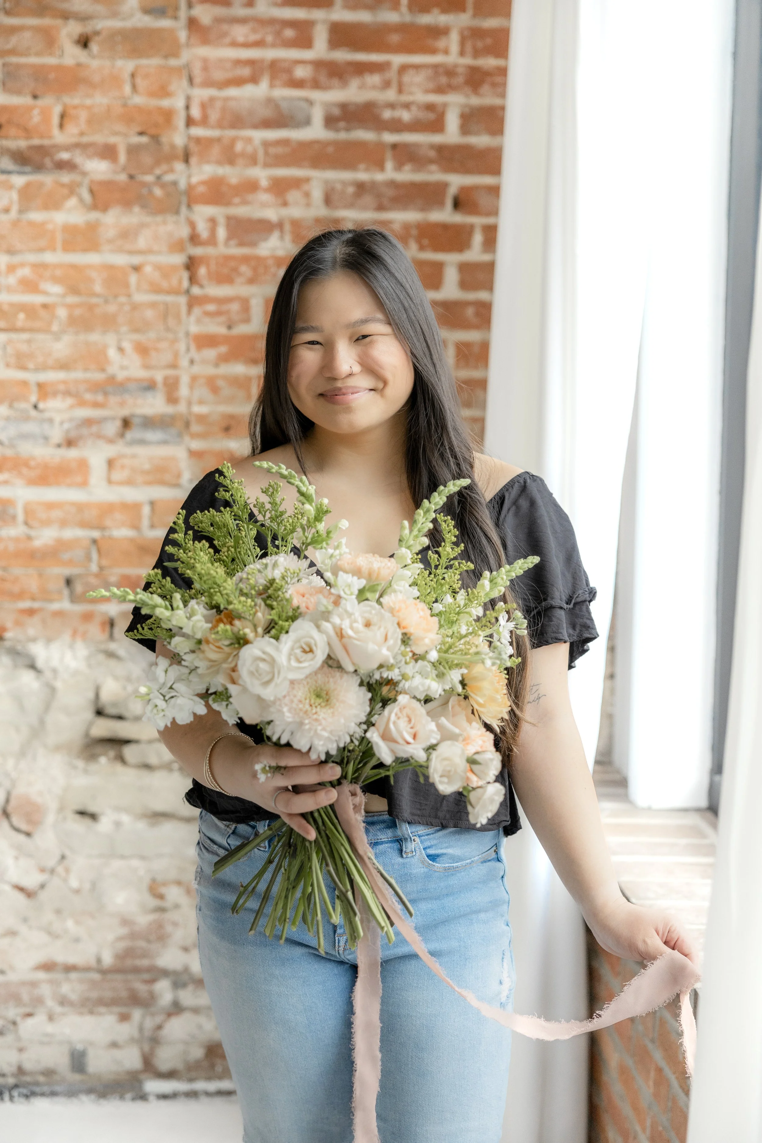 A woman with long dark hair holding a bouquet of white and peach flowers, standing in front of a brick wall and white curtains.