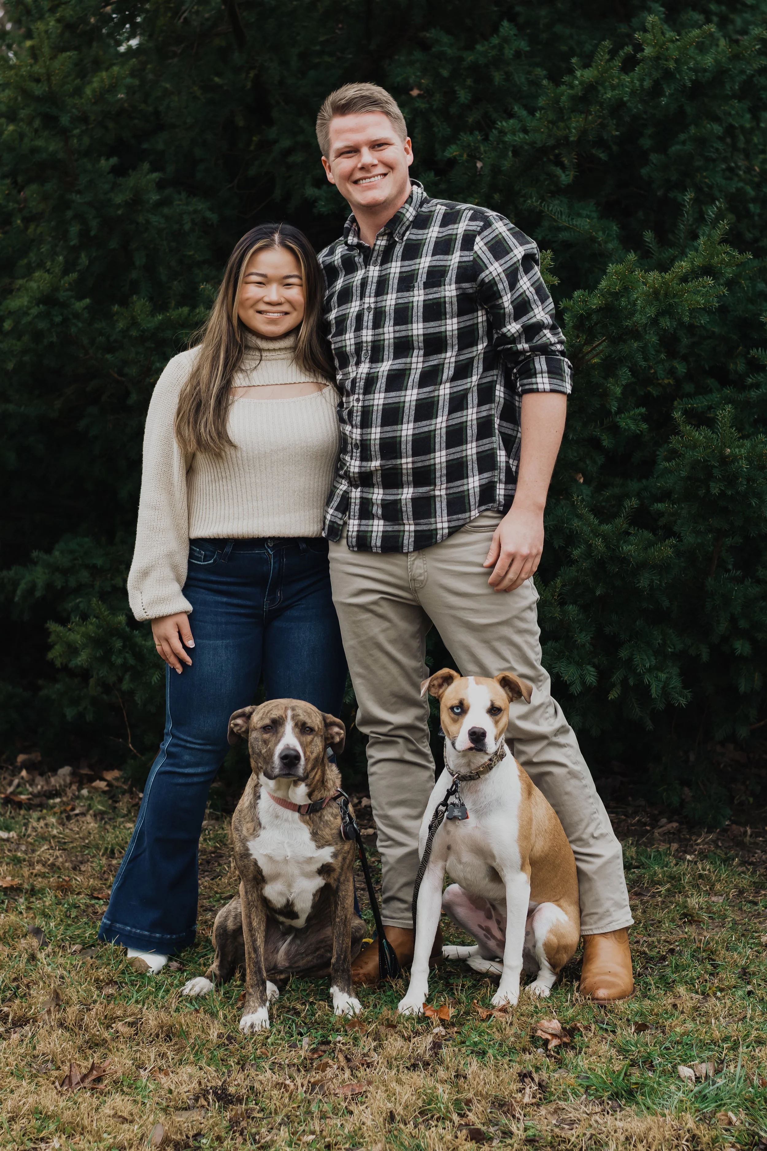 A smiling couple standing outdoors in front of green bushes, with two dogs sitting at their feet.