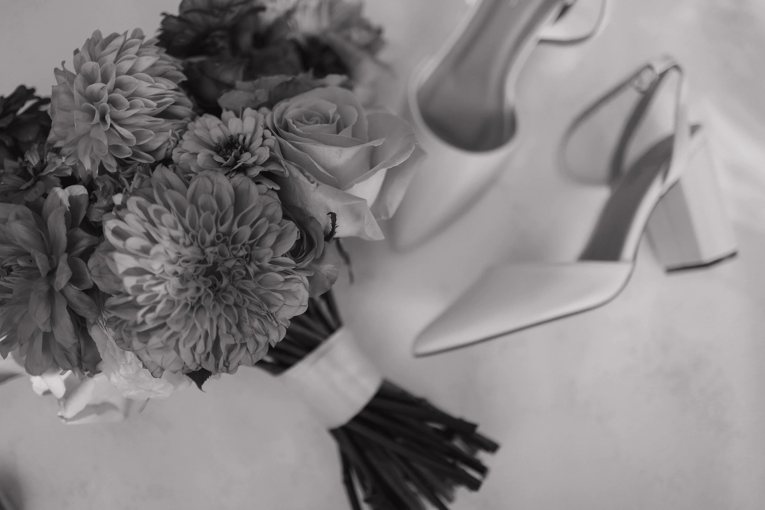 A black and white photo of a bouquet of flowers, including roses and dahlias, placed on a surface near a pair of high-heeled shoes.