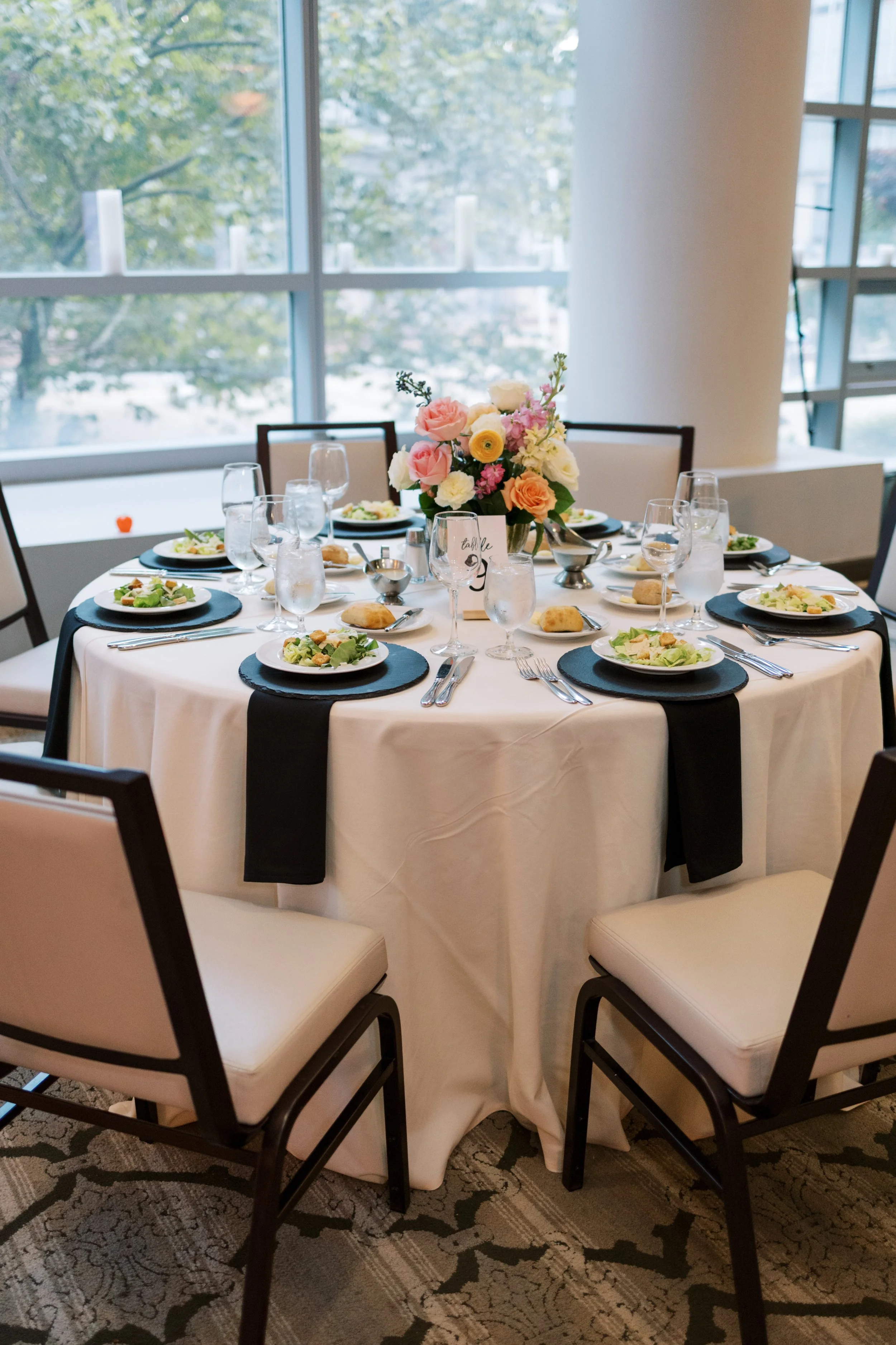 Round banquet table set with black placemats, white napkins, silverware, glasses, a floral centerpiece, and plates of salad, arranged for an event in a room with large windows.
