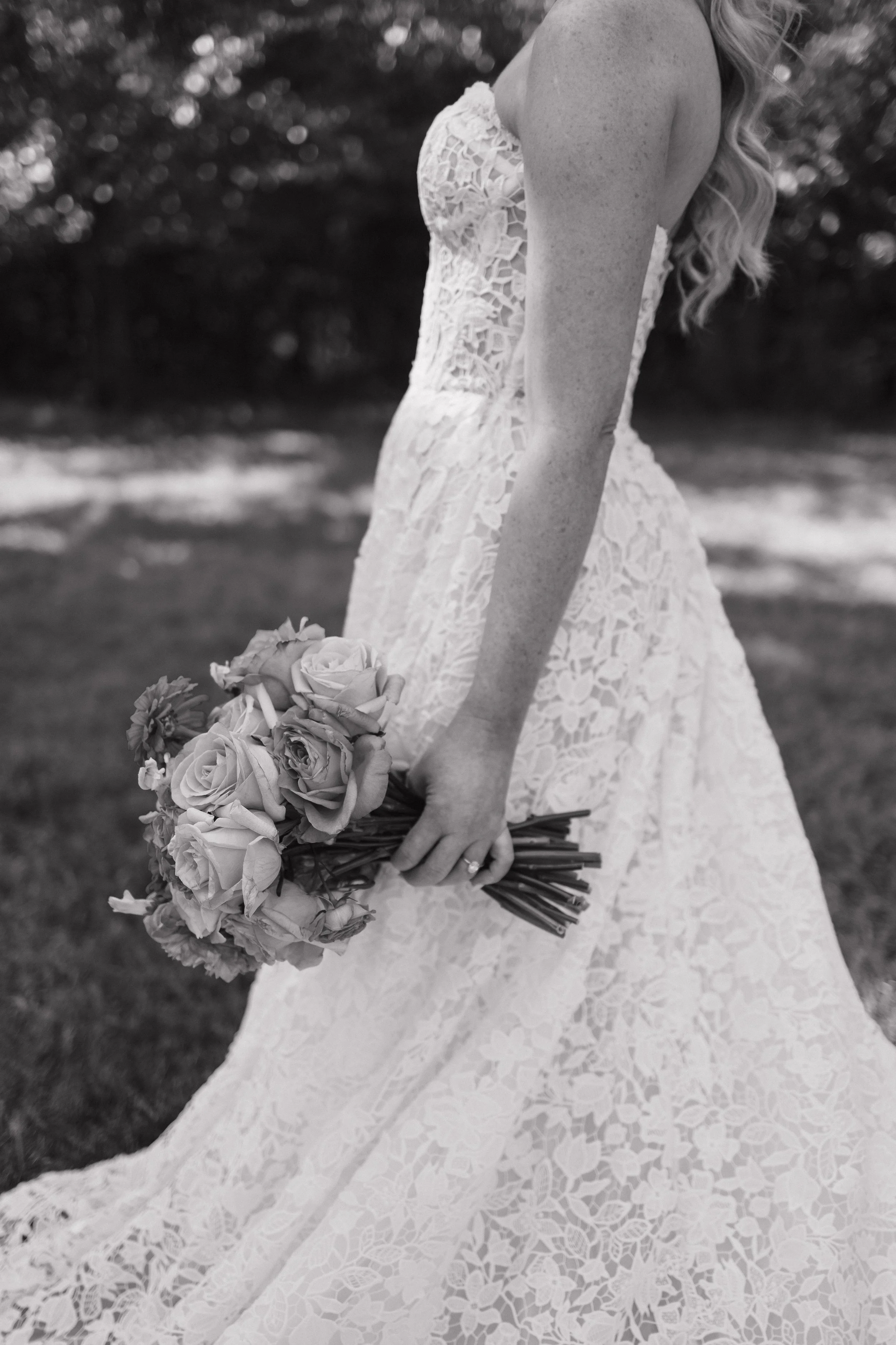 A woman in a lace wedding dress holding a bouquet of flowers outdoors.