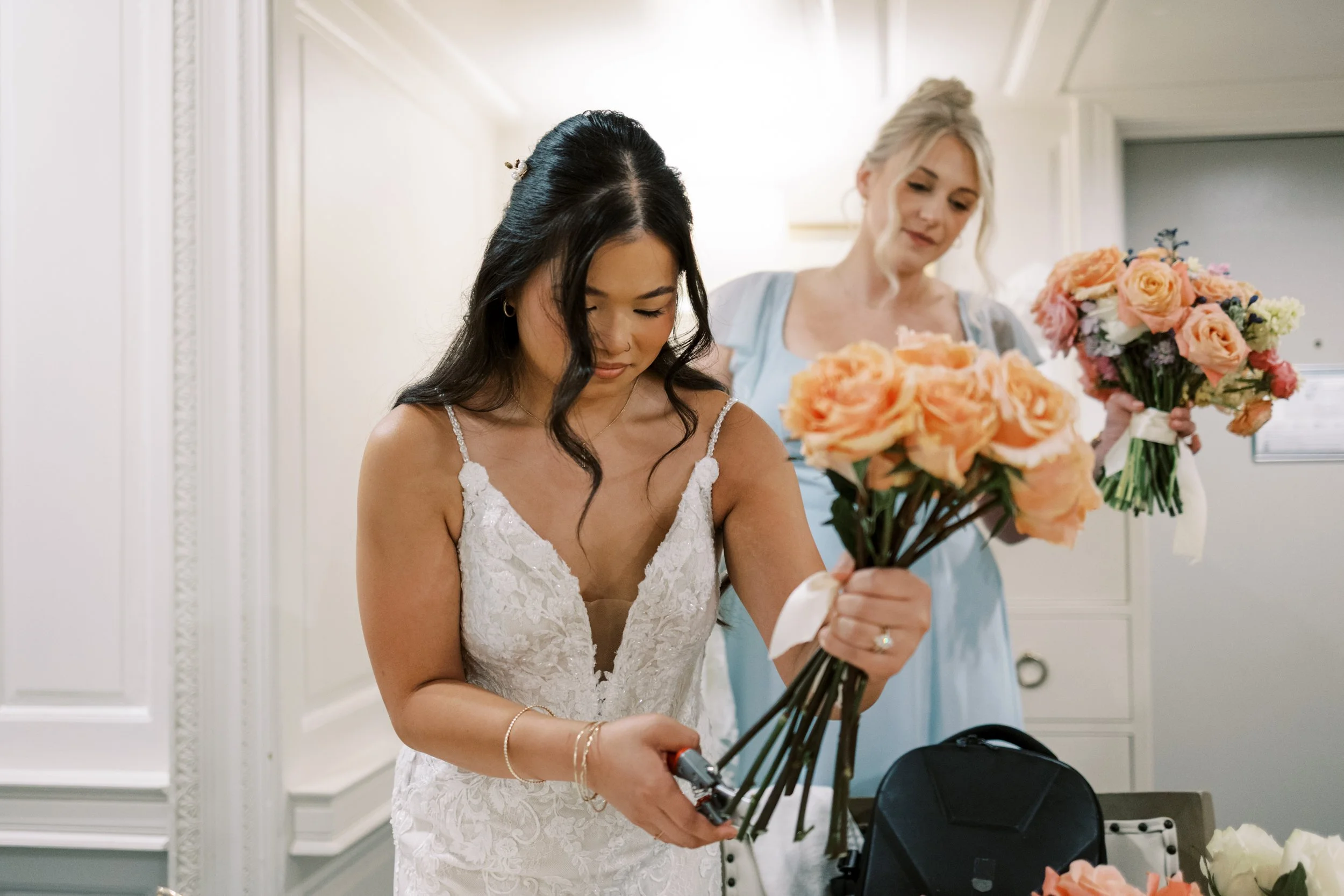 Bride in a white lace dress holding a bouquet of peach roses, with a woman in a light blue dress and another person holding a similar bouquet in the background.