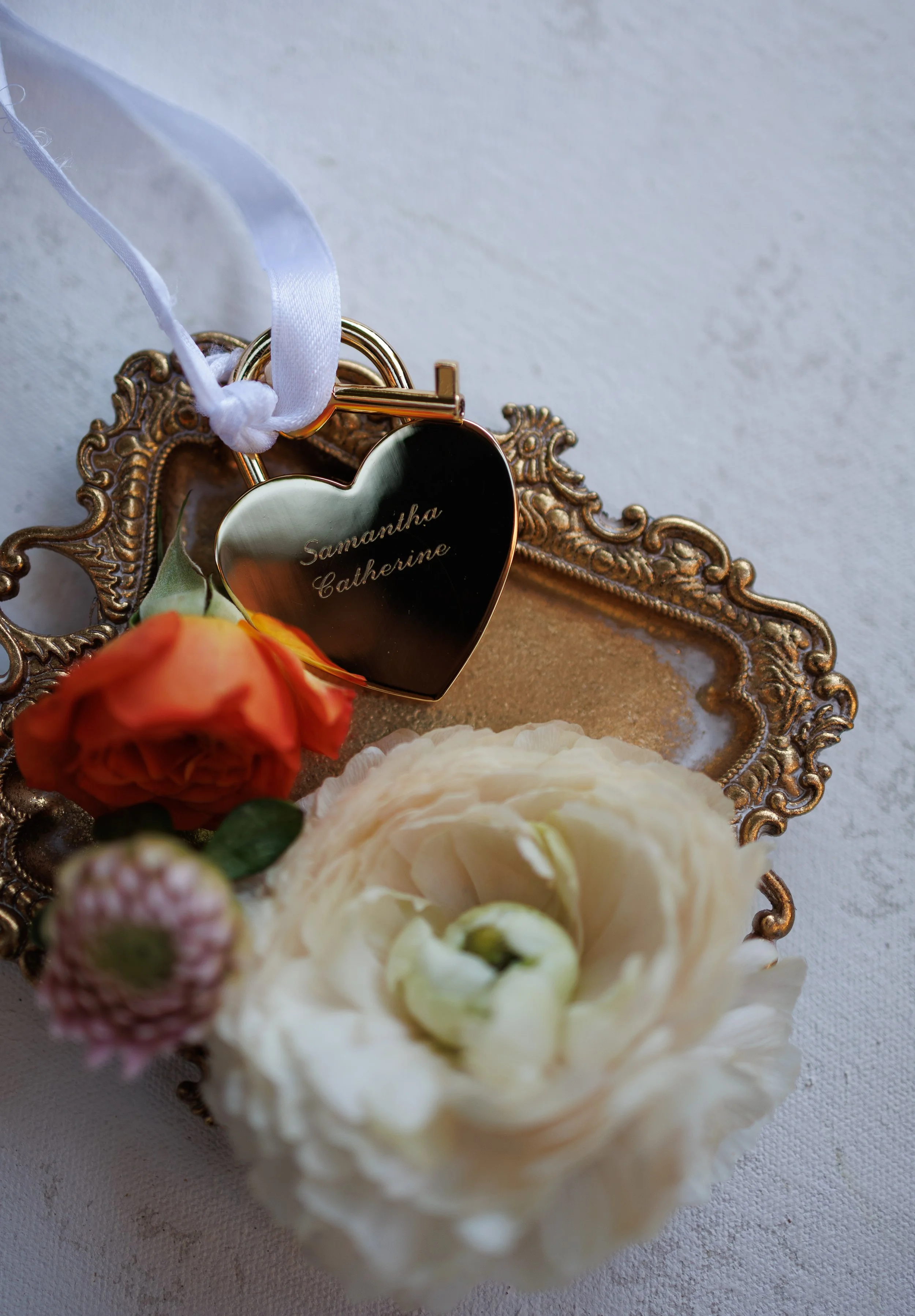 A decorative display with a gold heart-shaped lock engraved with the names "Samantha Catherine," a white ribbon, a peach-colored rose, a pinkish flower, and a white ranunculus flower arranged on an ornate gold tray with a white textured surface background.