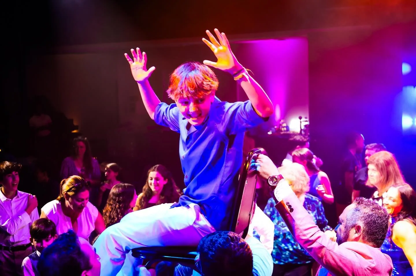 A young man dancing on someone's lap during a lively party with colorful lighting and smiling crowd.