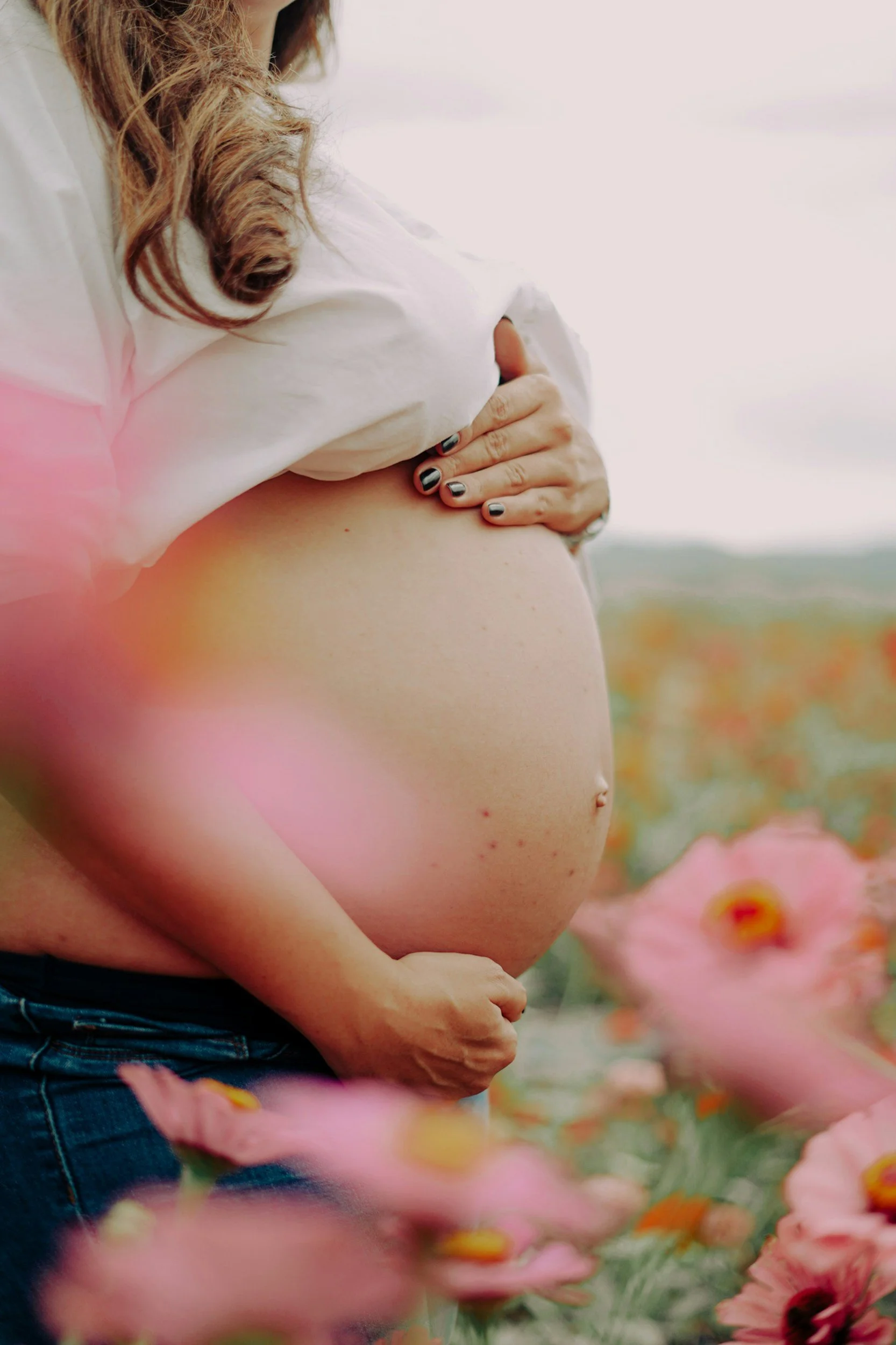 A pregnant woman with long hair holding her belly, standing in a field of pink flowers.