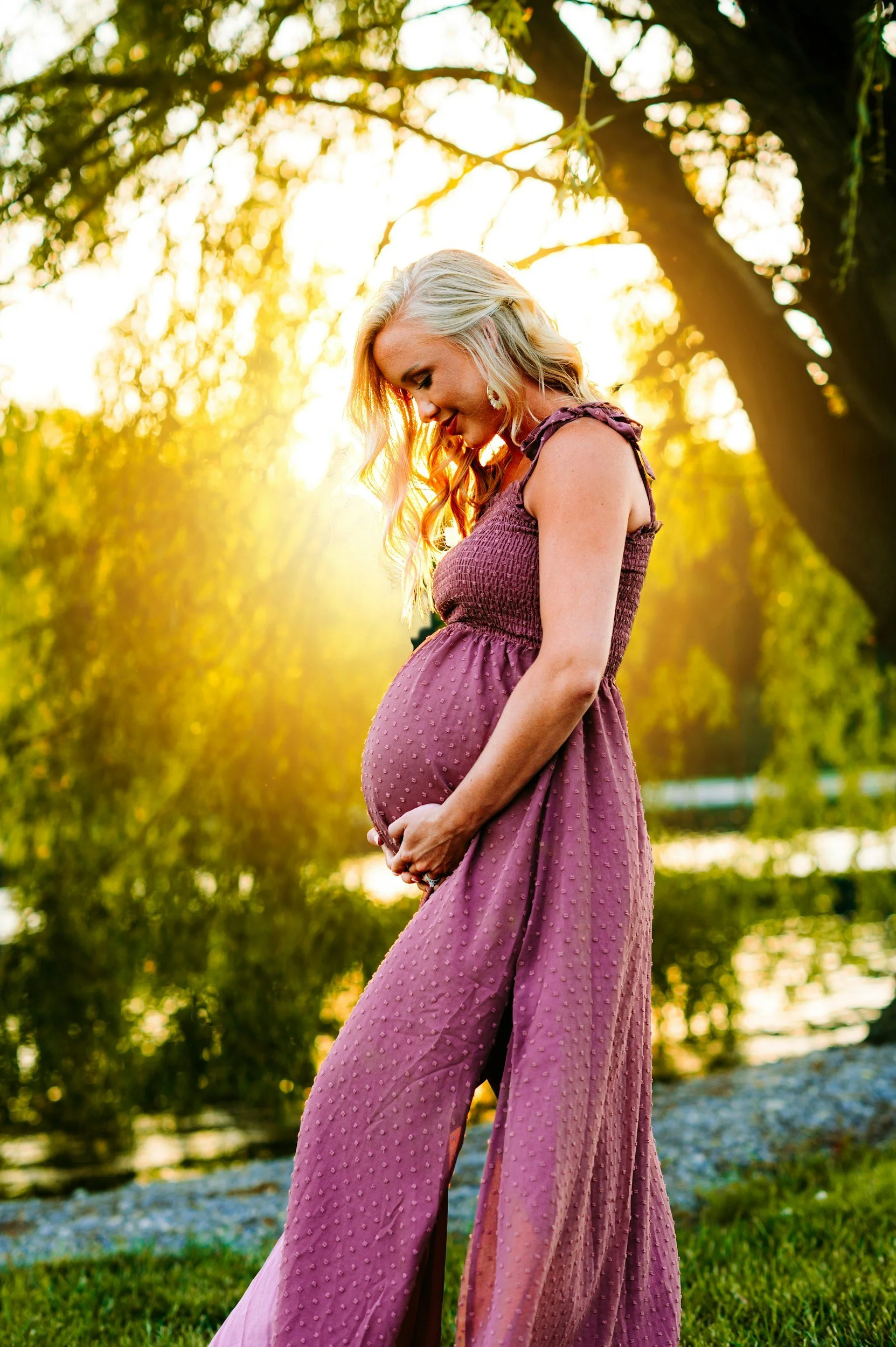 Pregnant woman wearing a mauve dress, standing outdoors in a natural setting with sunlight filtering through trees, looking down and holding her belly.