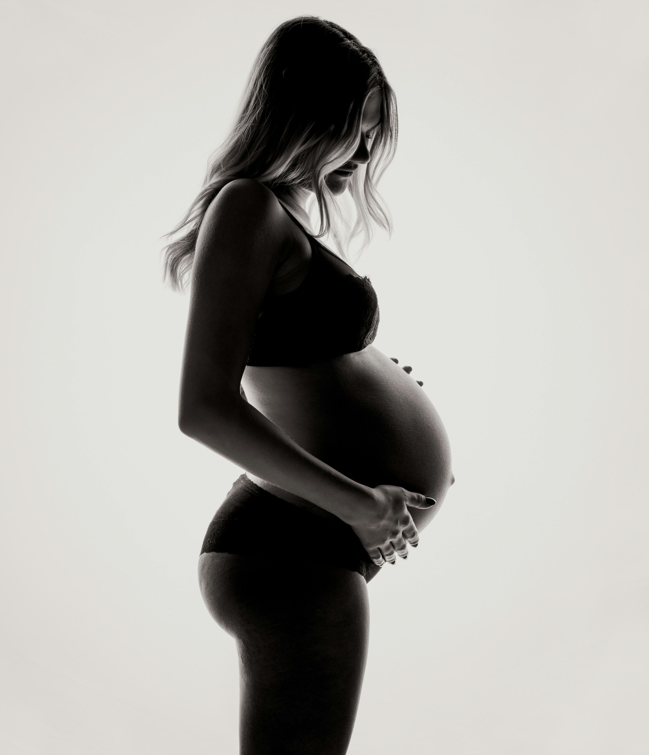 Silhouette of a pregnant woman wearing underwear and a bra, gently holding her belly, in black and white