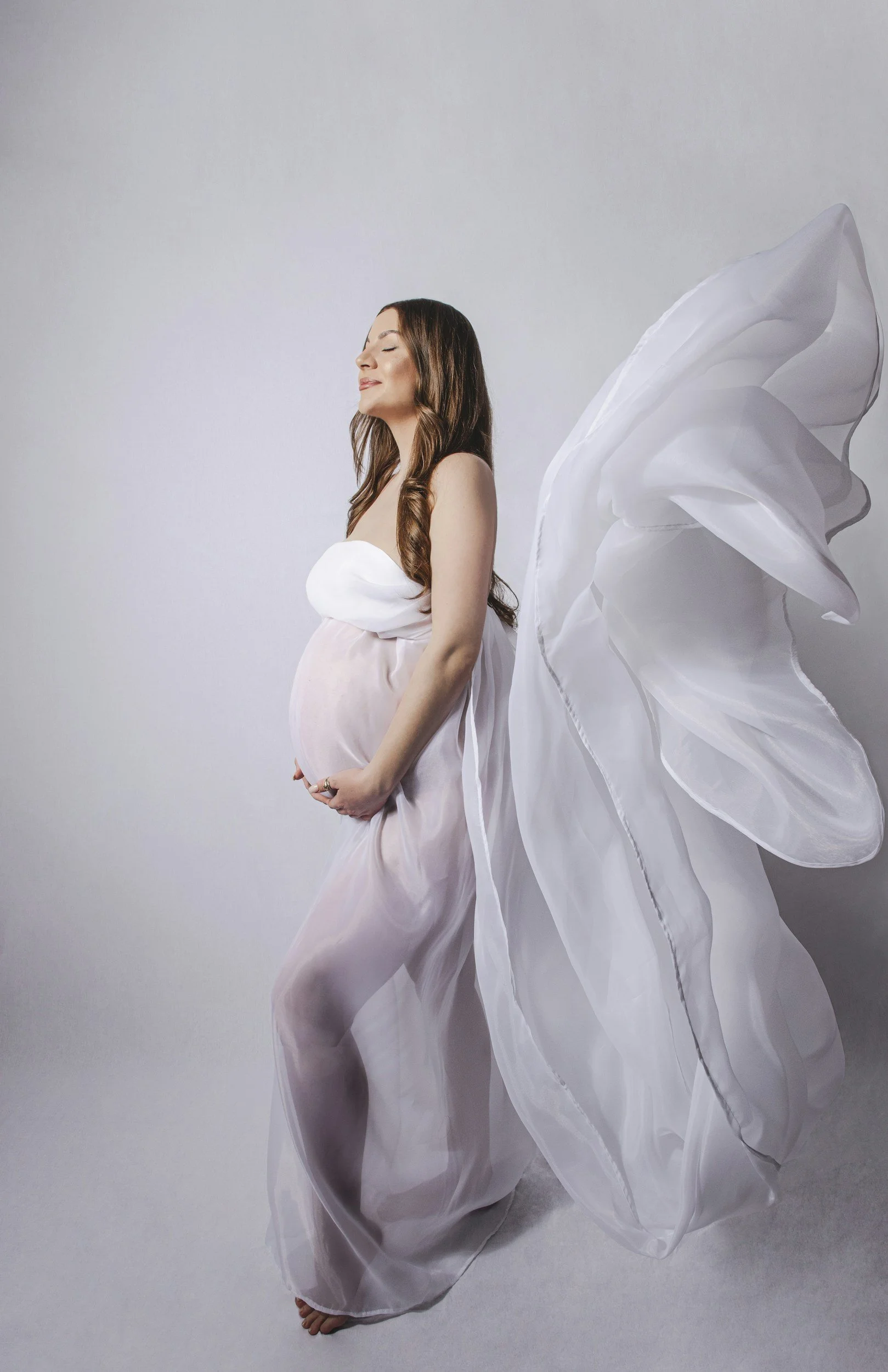 Pregnant woman with long brown hair in a flowing dress with angel wings.