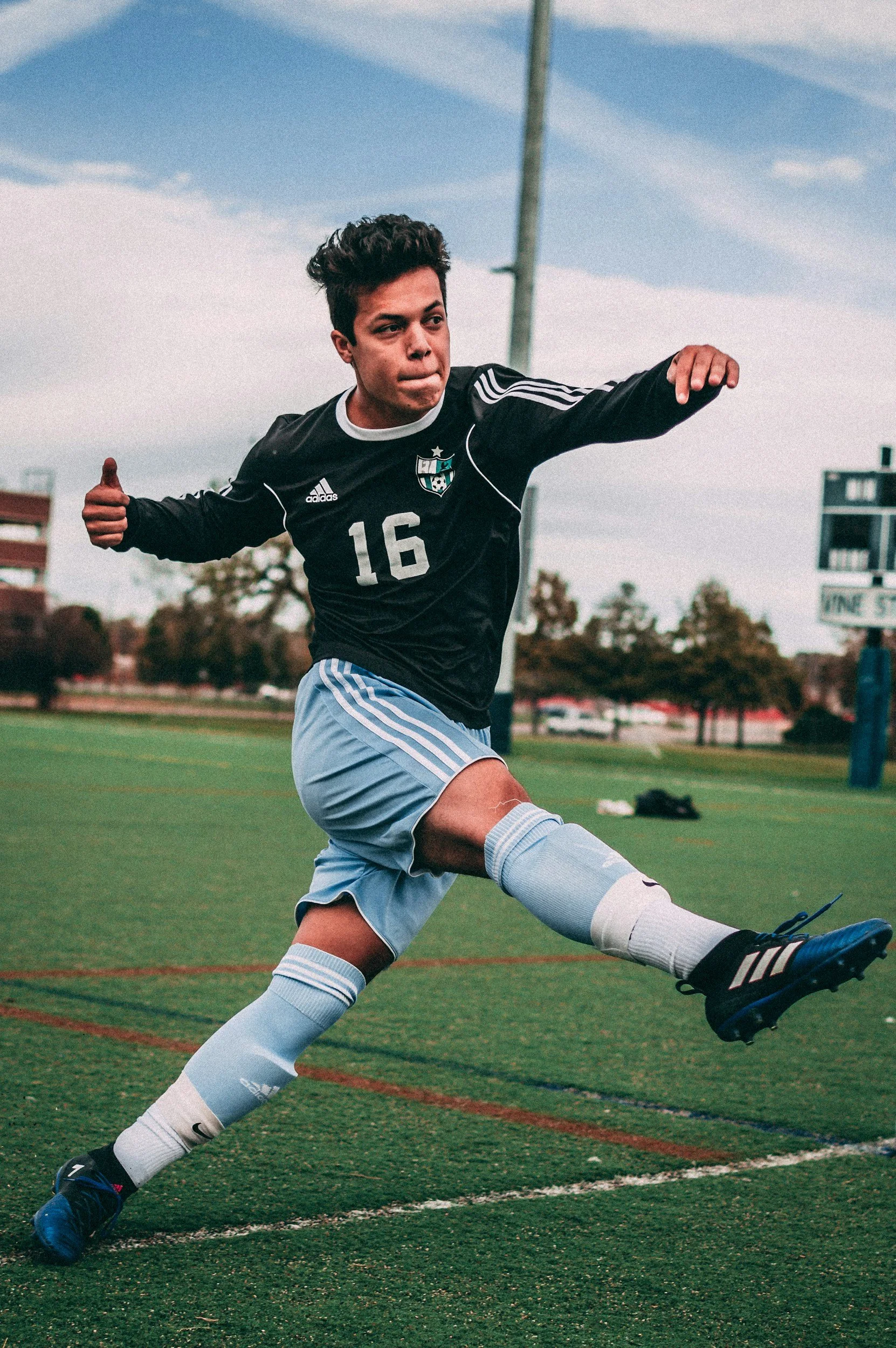 A young male soccer player in a black jersey with the number 16, gray shorts, and blue cleats is mid-kick on a green field with trees and a building in the background.