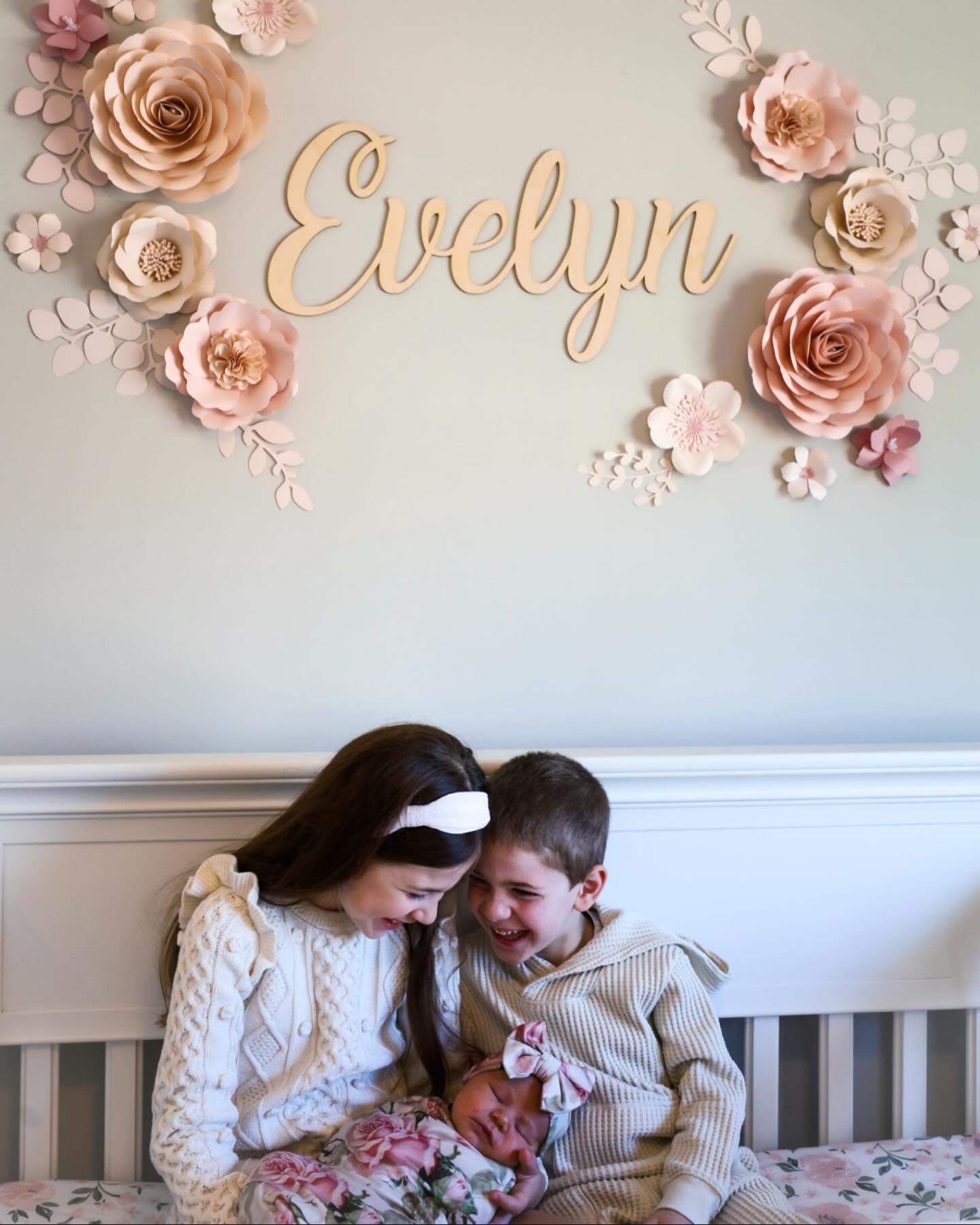 Two children, a girl and a boy, sitting on a bed with their newborn sister. The girl and boy are smiling and looking at their sister, who is sleeping. The wall behind them has paper flower decorations and the name "Evelyn" in cursive, with pink and white flowers around.