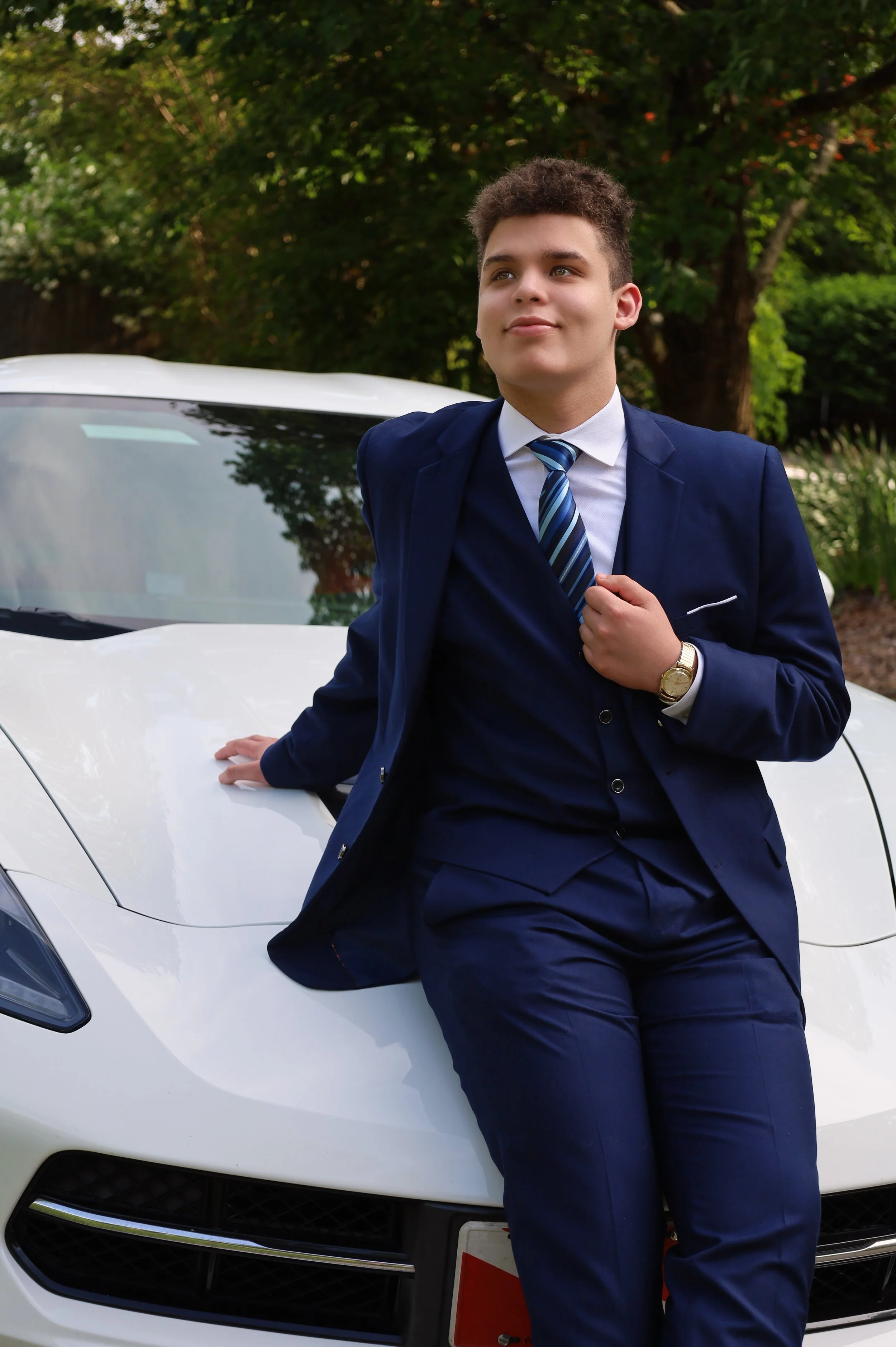 A young man dressed in a navy suit and striped tie leaning against the front of a white car, with trees and greenery in the background.