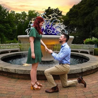 A man on one knee proposing to a woman in front of a decorative fountain at sunset.