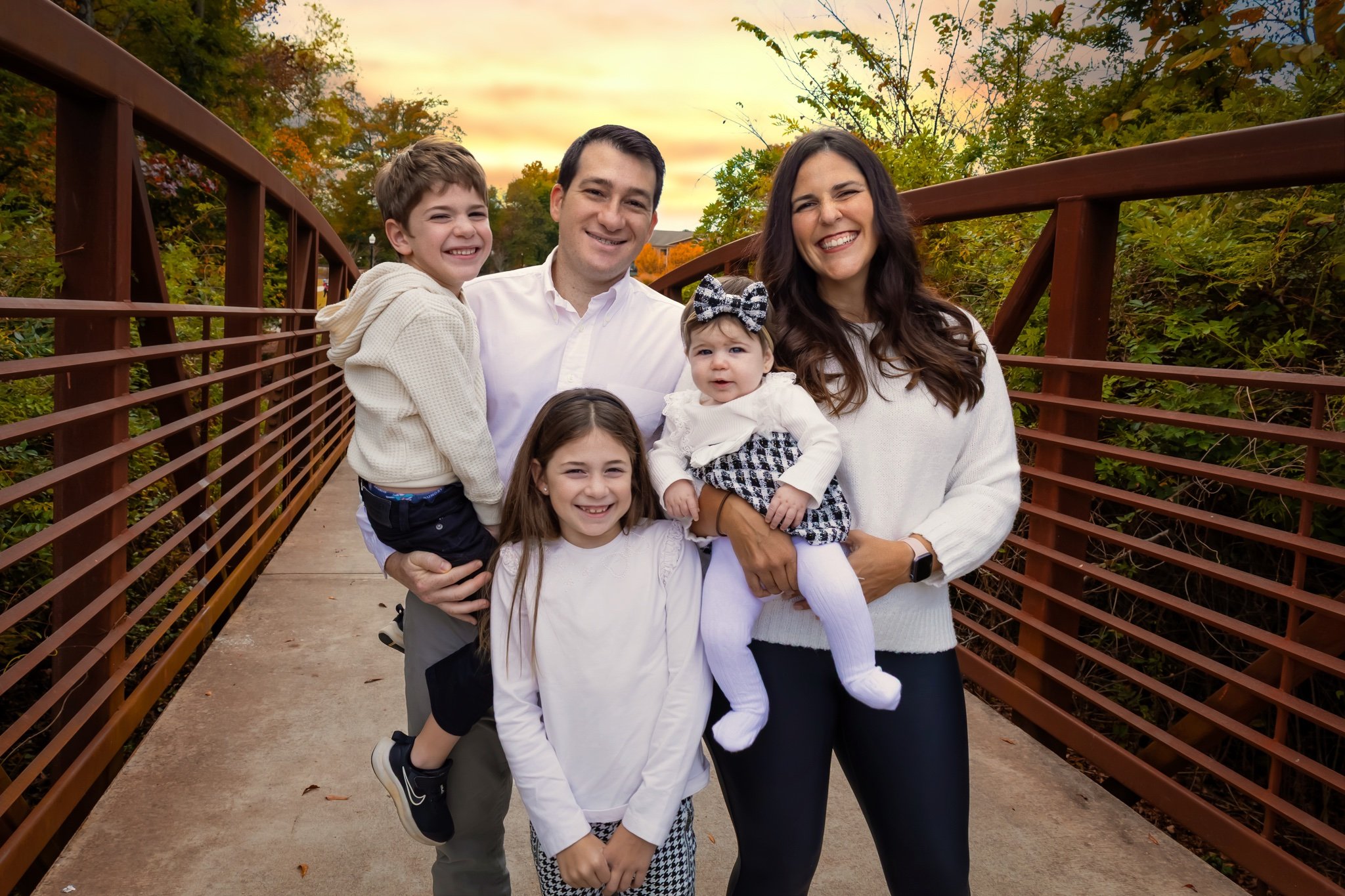 Family of six posing on a bridge in a park during autumn sunset, smiling and dressed in white and neutral clothing.