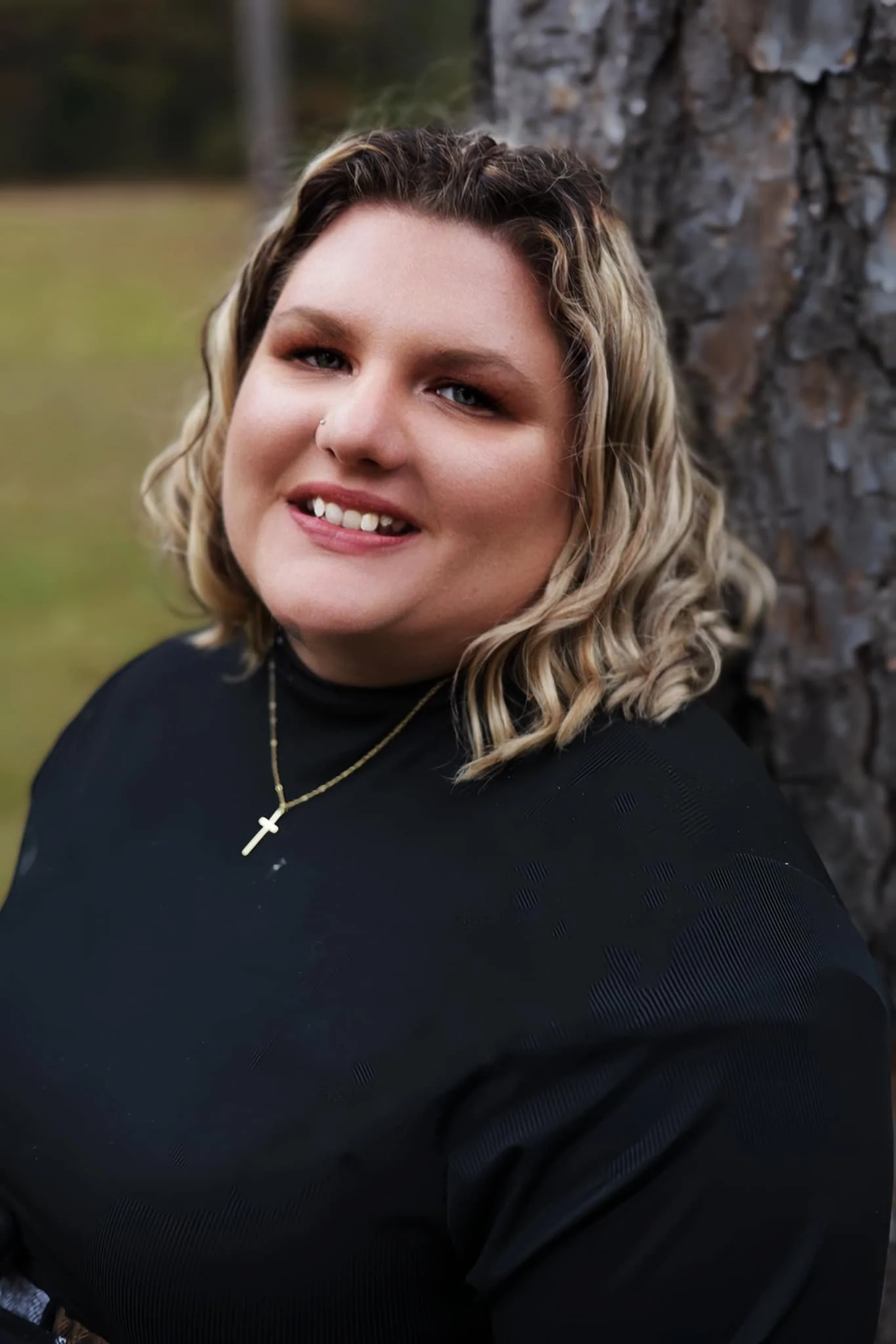 A smiling woman with blonde, shoulder-length curly hair, wearing a black top and a gold cross necklace, leaning against a tree outdoors with a blurred natural background.